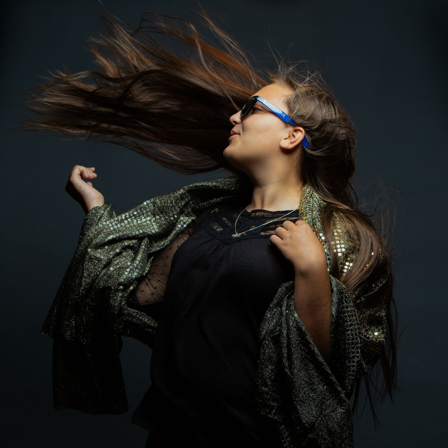 Woman with flowing hair wearing blue glasses and a shimmering green jacket poses dynamically against a dark background.