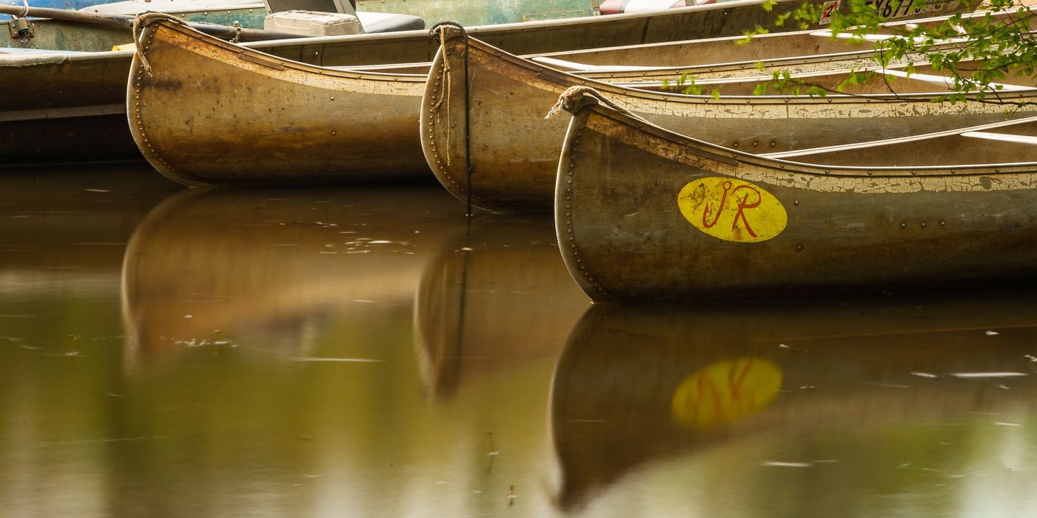 Weathered wooden canoes with yellow labels rest on calm reflective water at a dock.