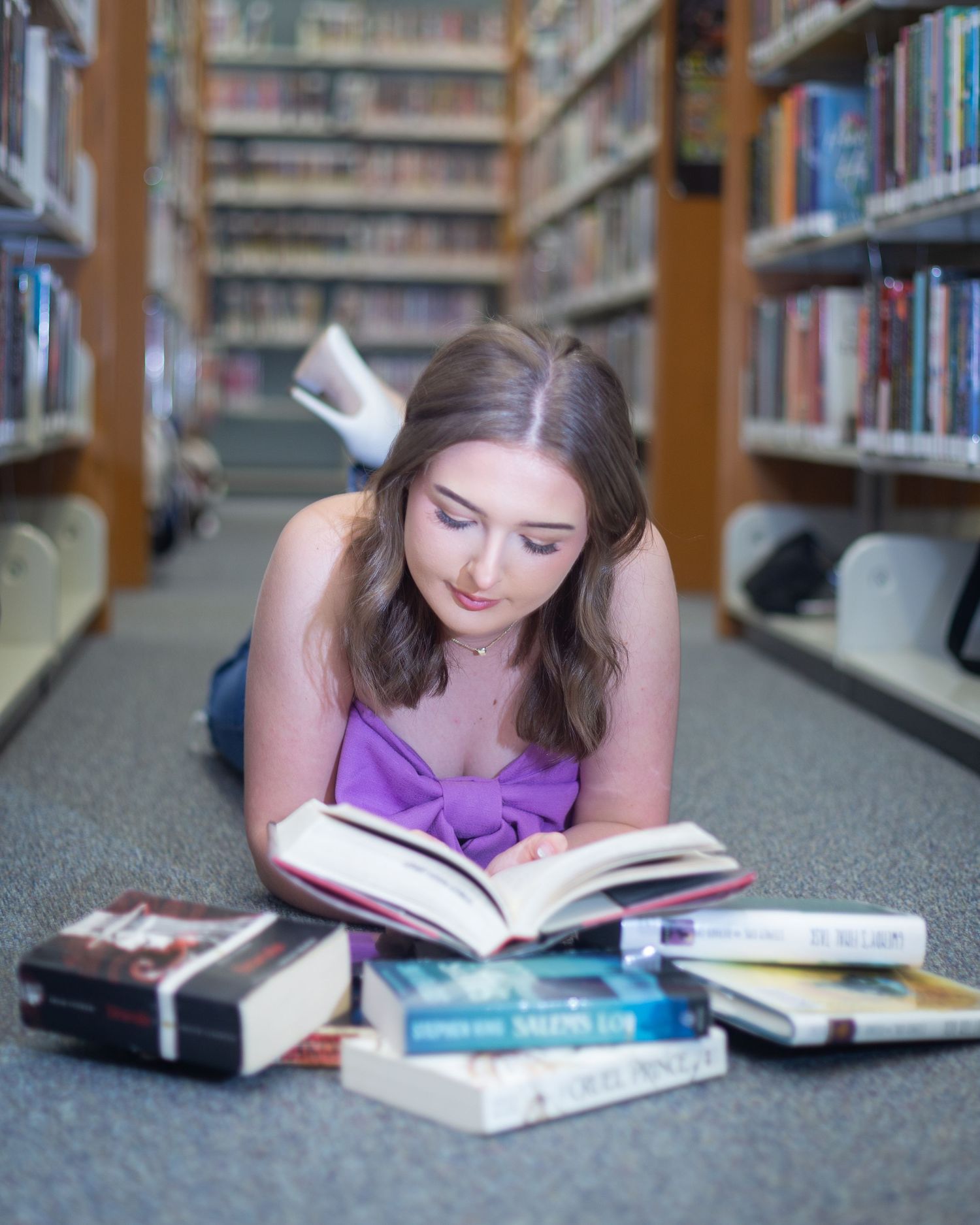 Student lying on library floor surrounded by open books while studying in between bookshelves.