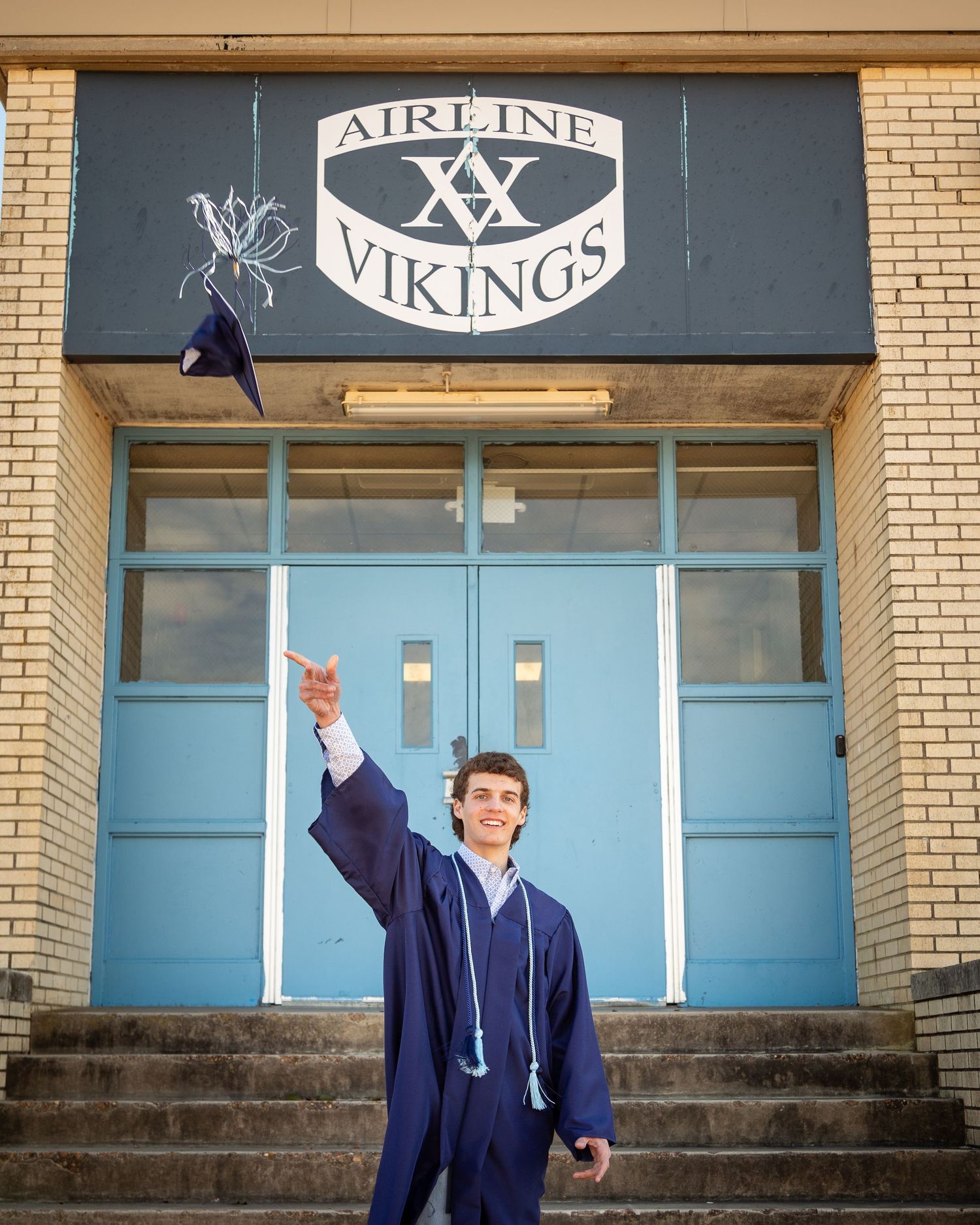 Graduate in navy blue cap and gown poses on brick building steps with raised arms in celebration.