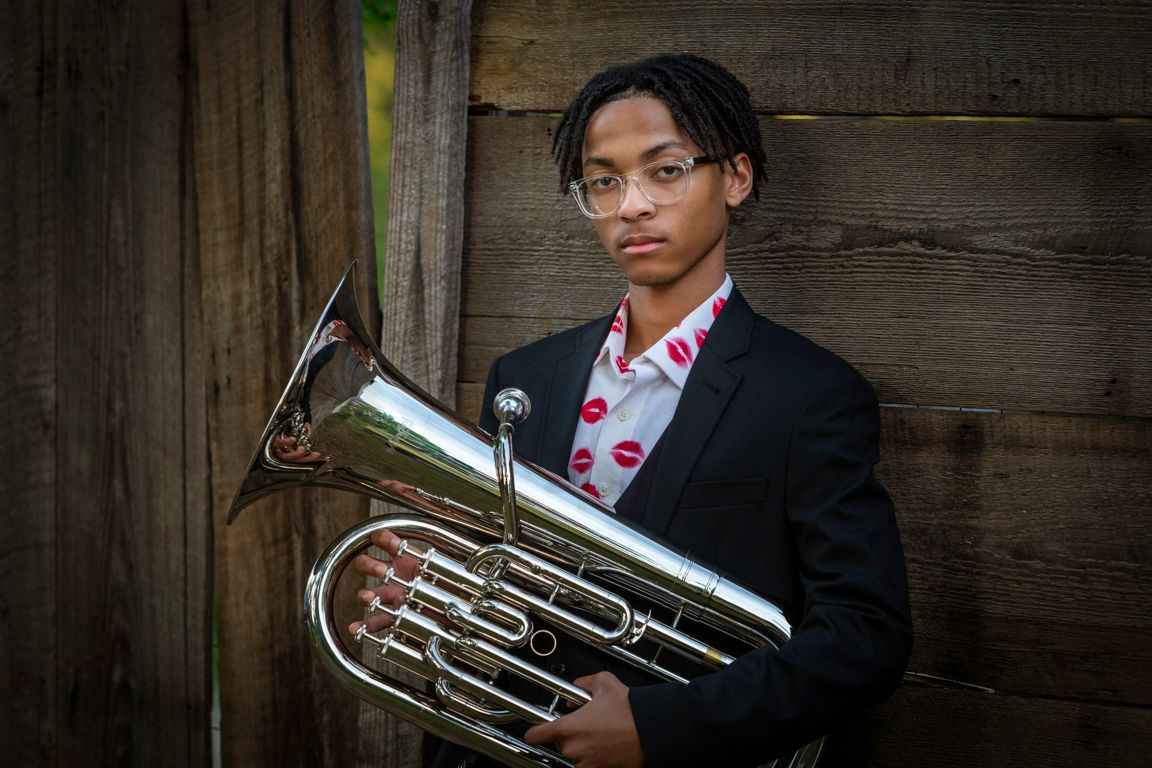 A high school senior musician in formal attire holds a brass musical instrument against a rustic wooden wall background.