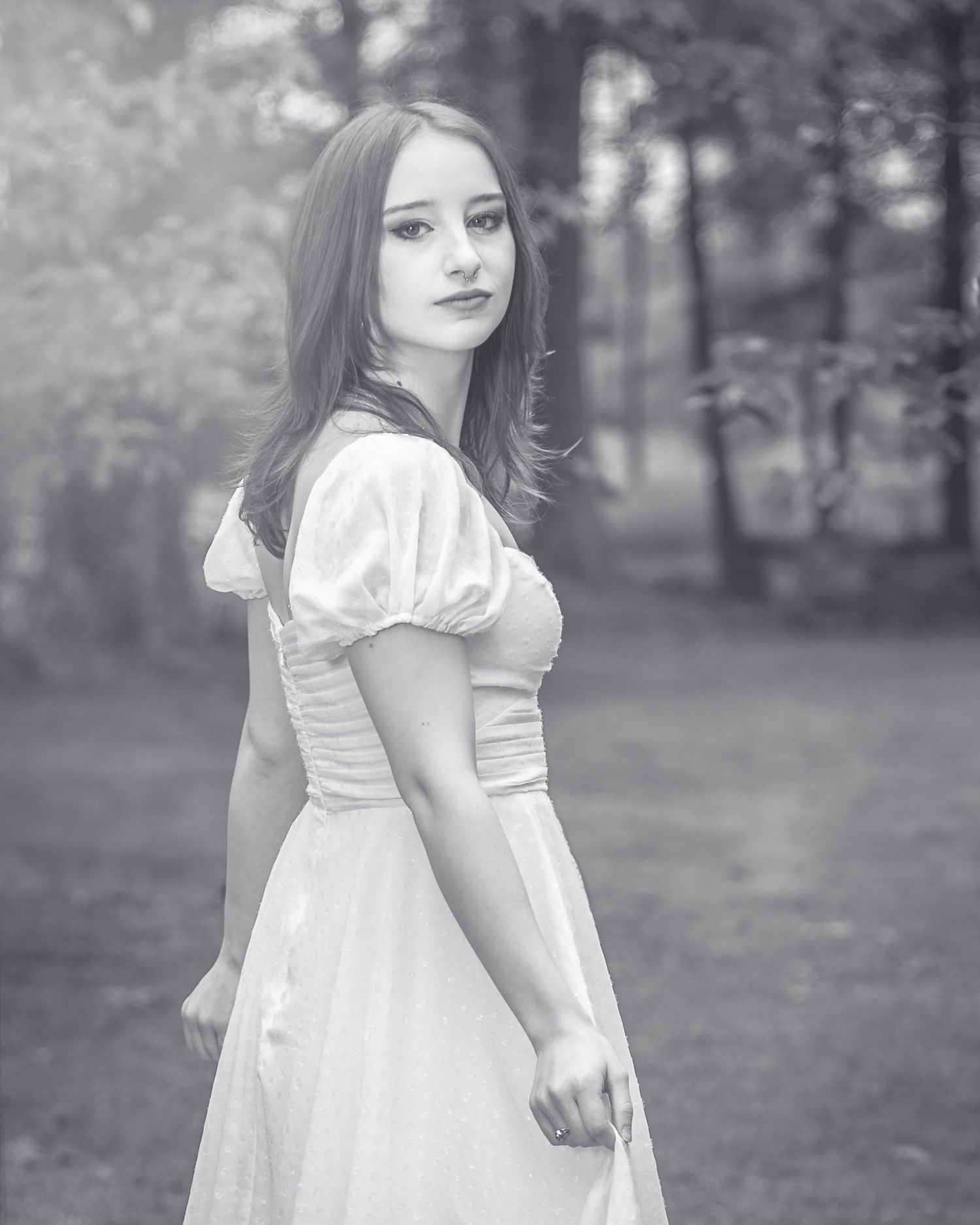 A young girl in a white dress twirls and plays in a wooded outdoor setting, creating a dreamy black and white scene.