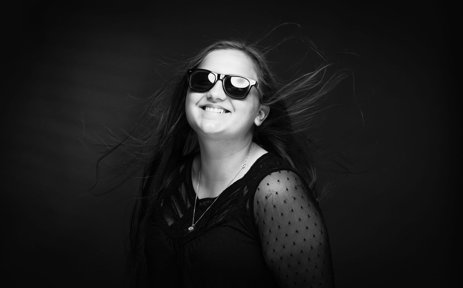 Black and white portrait of a smiling woman with sunglasses, hair blowing in the wind against a dark background.