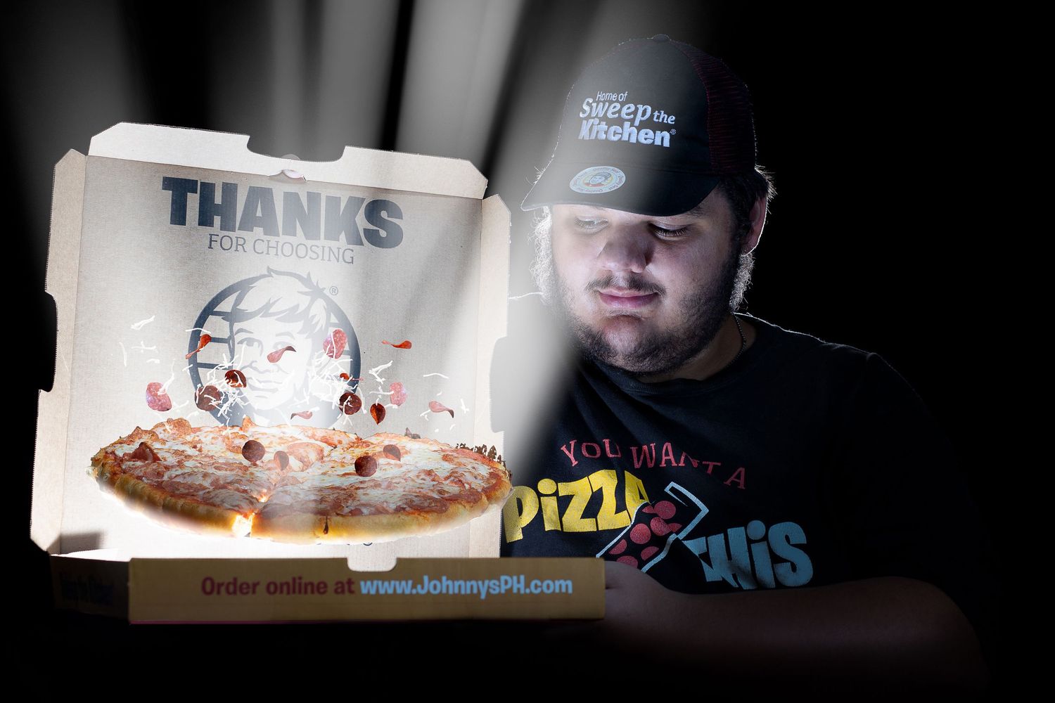 Man in baseball cap and pizza shirt poses beside an open pizza box with a thank you message inside.