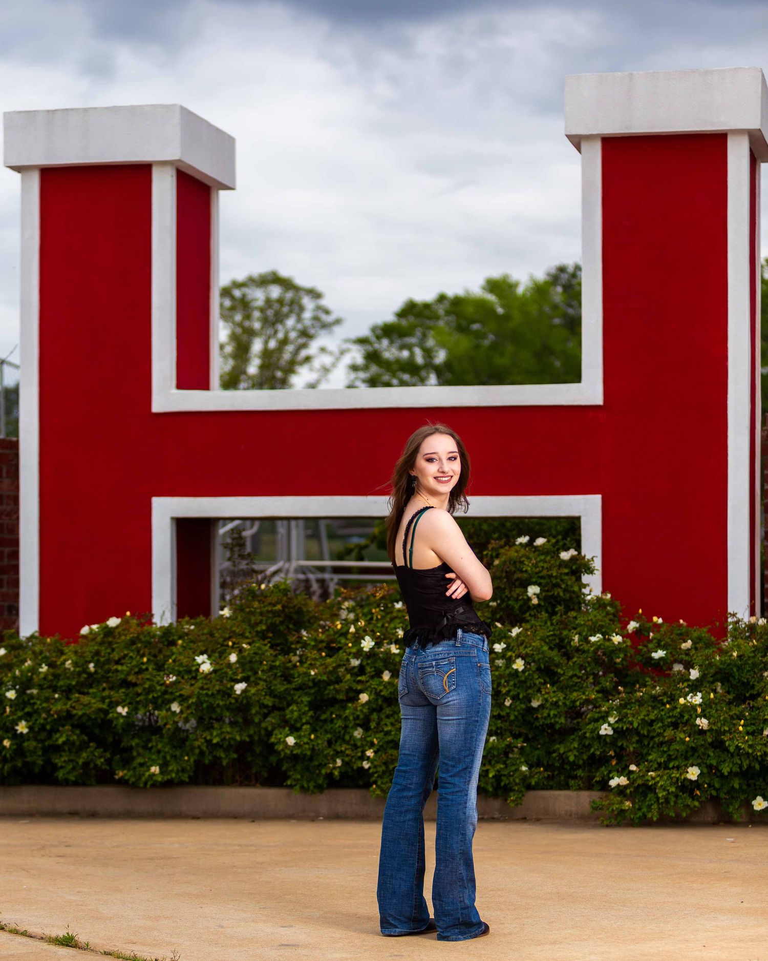 A high school senior in blue jeans poses against a modern red and white architectural backdrop with shrubs and trees.