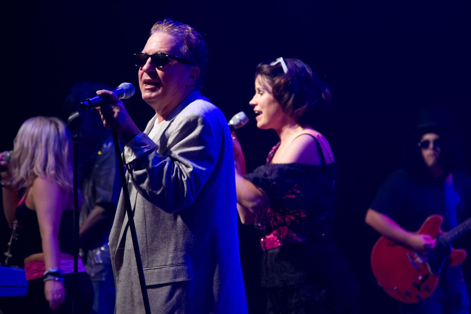 Two singers perform on stage with microphones, backed by band members with guitars in a dimly lit concert venue.