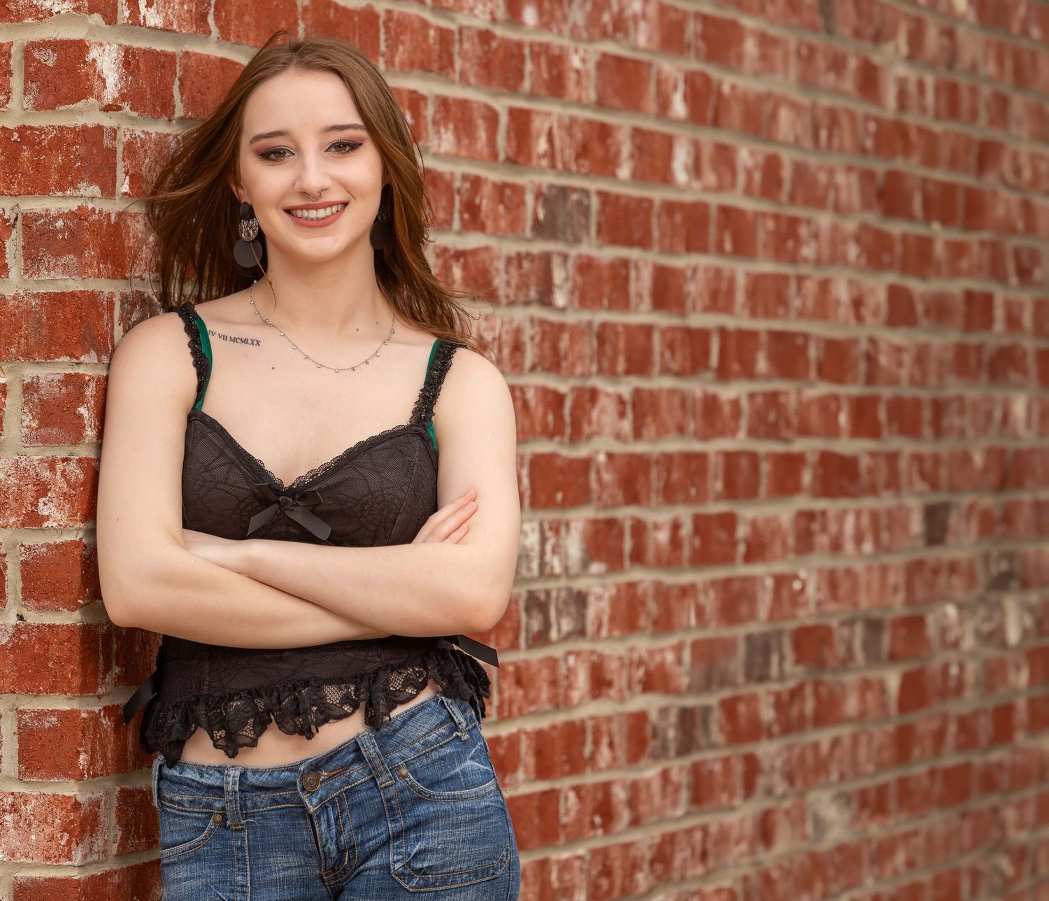 A person in a black lace top and jeans poses casually against a rustic brick wall.