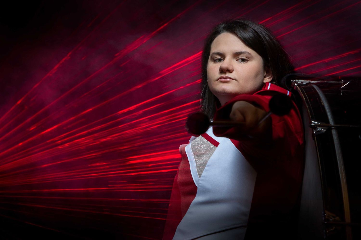 A high school student  in a marching band uniformt sits in a dark room illuminated by dramatic red laser light beams.