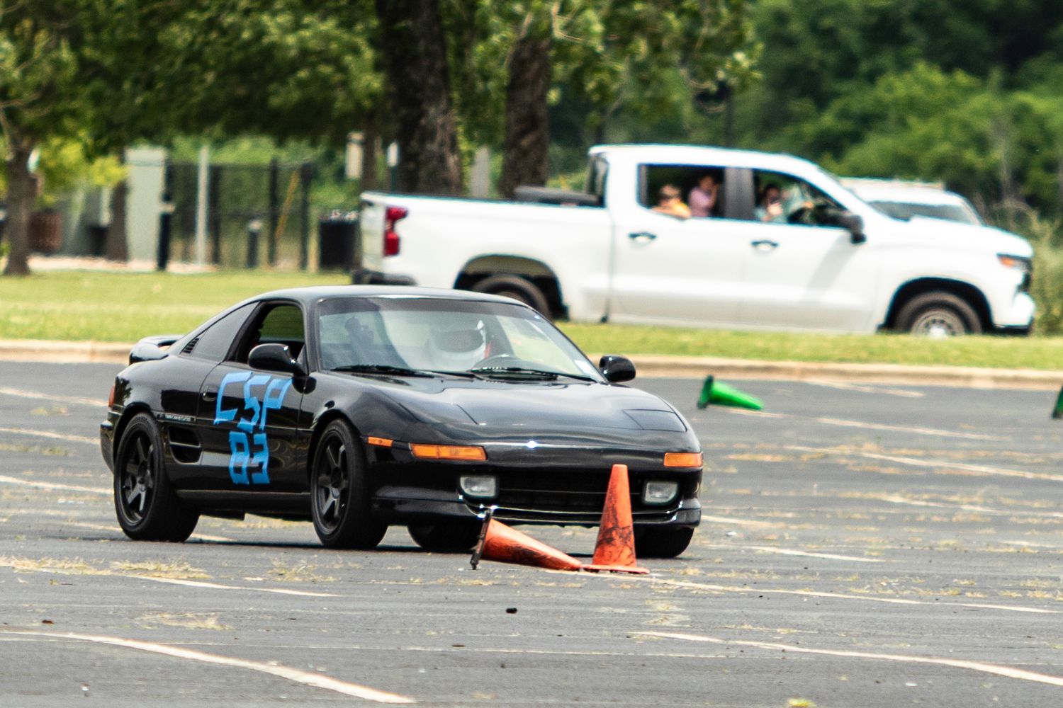 Black Toyota MR2 autocross racing car navigating orange cones on a parking lot course during a competitive event.