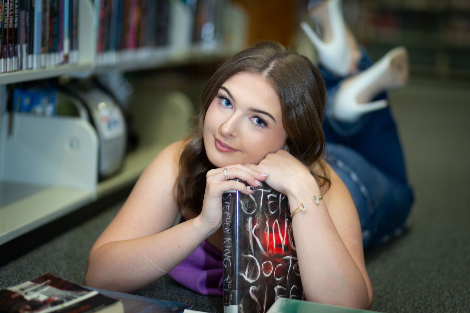 A student lies between bookshelves in a library while studying and posing for a photo.