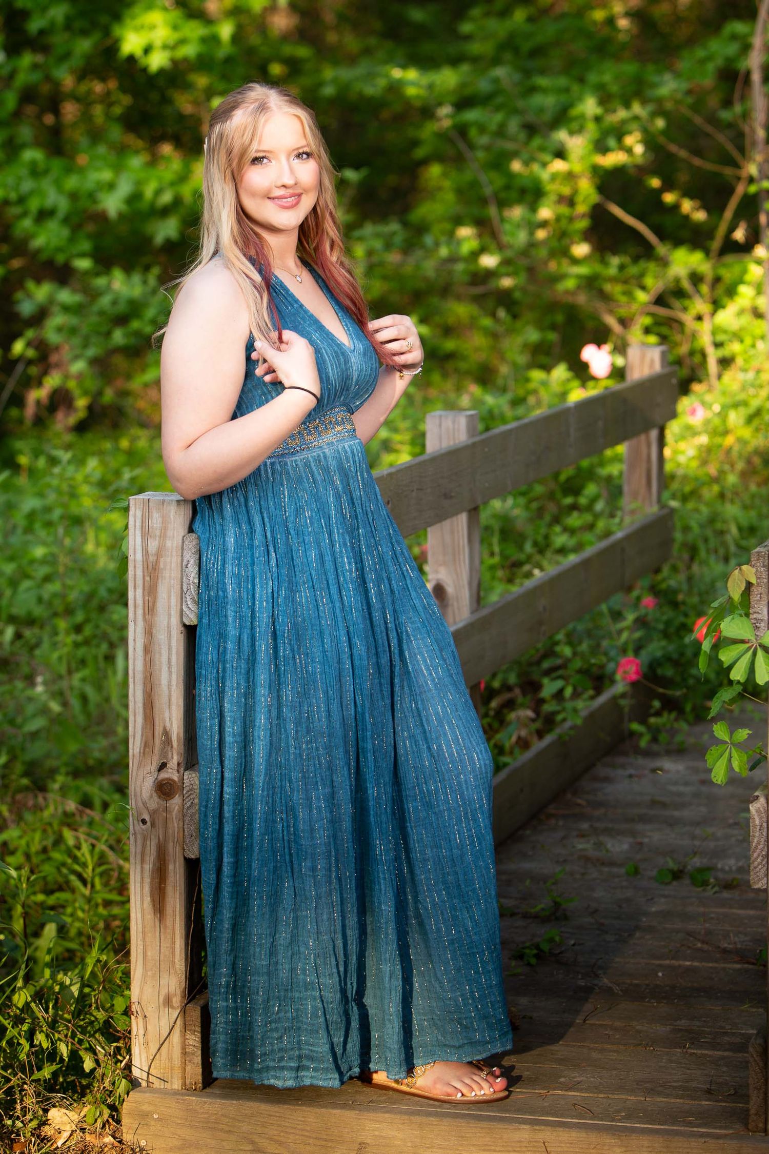 A person in a teal formal dress poses on a wooden walkway surrounded by lush green foliage.