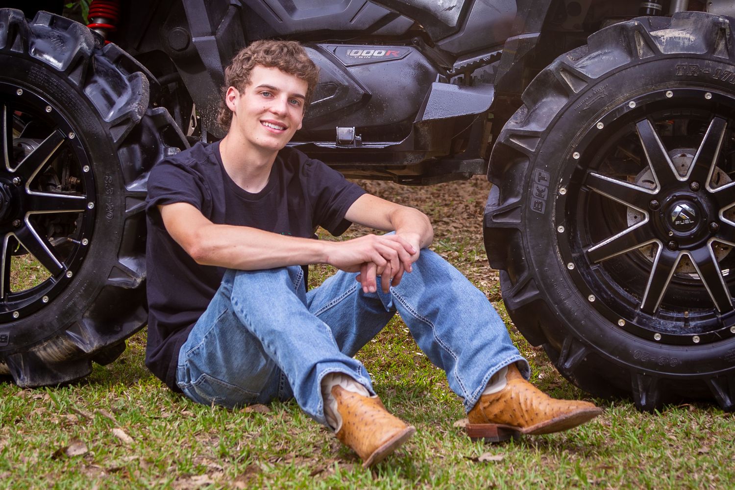 A person in jeans and boots sits casually on grass next to black motorcycle wheels and tires.