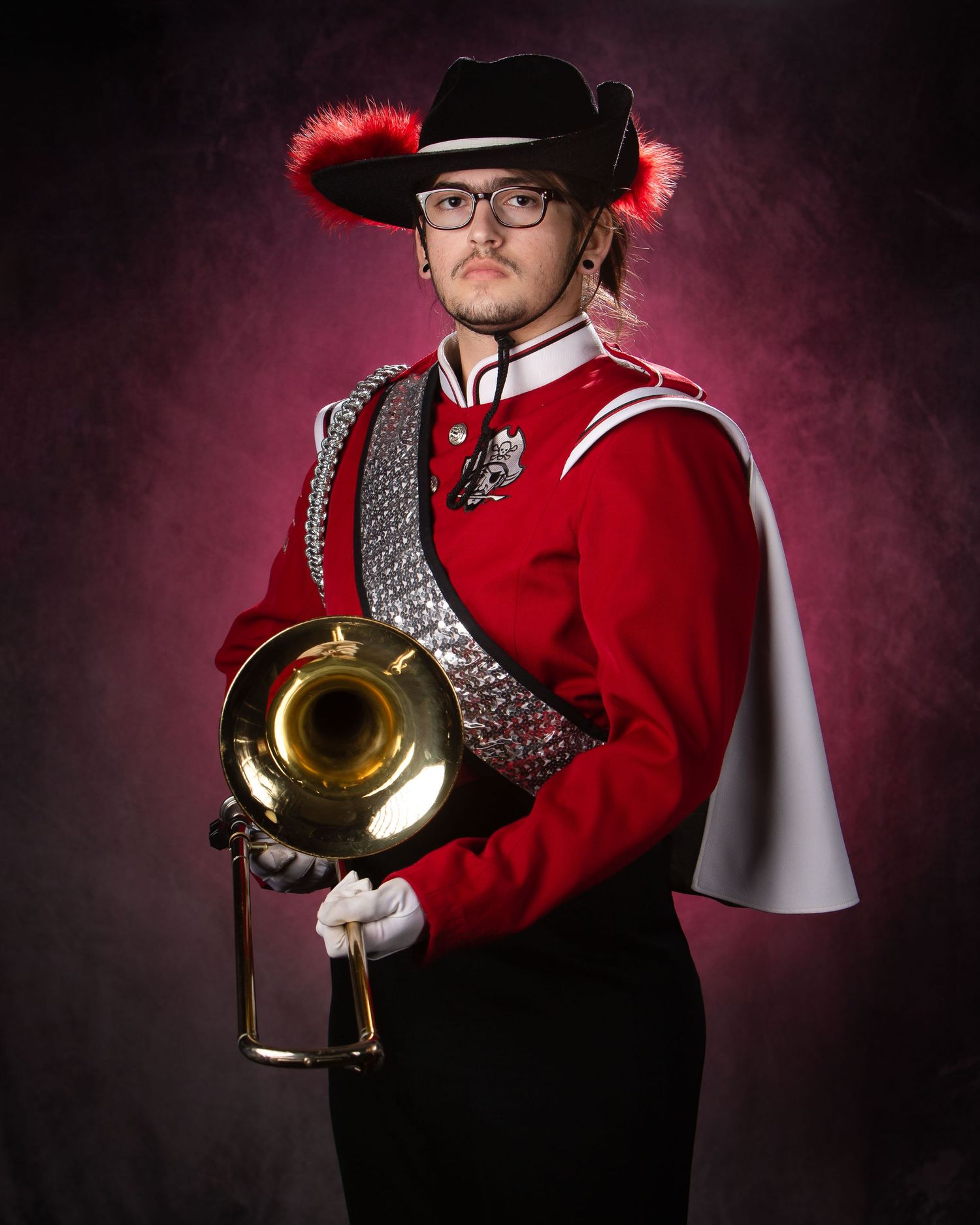 Musician in red marching band uniform holds brass trumpet against dramatic burgundy background with moody lighting.