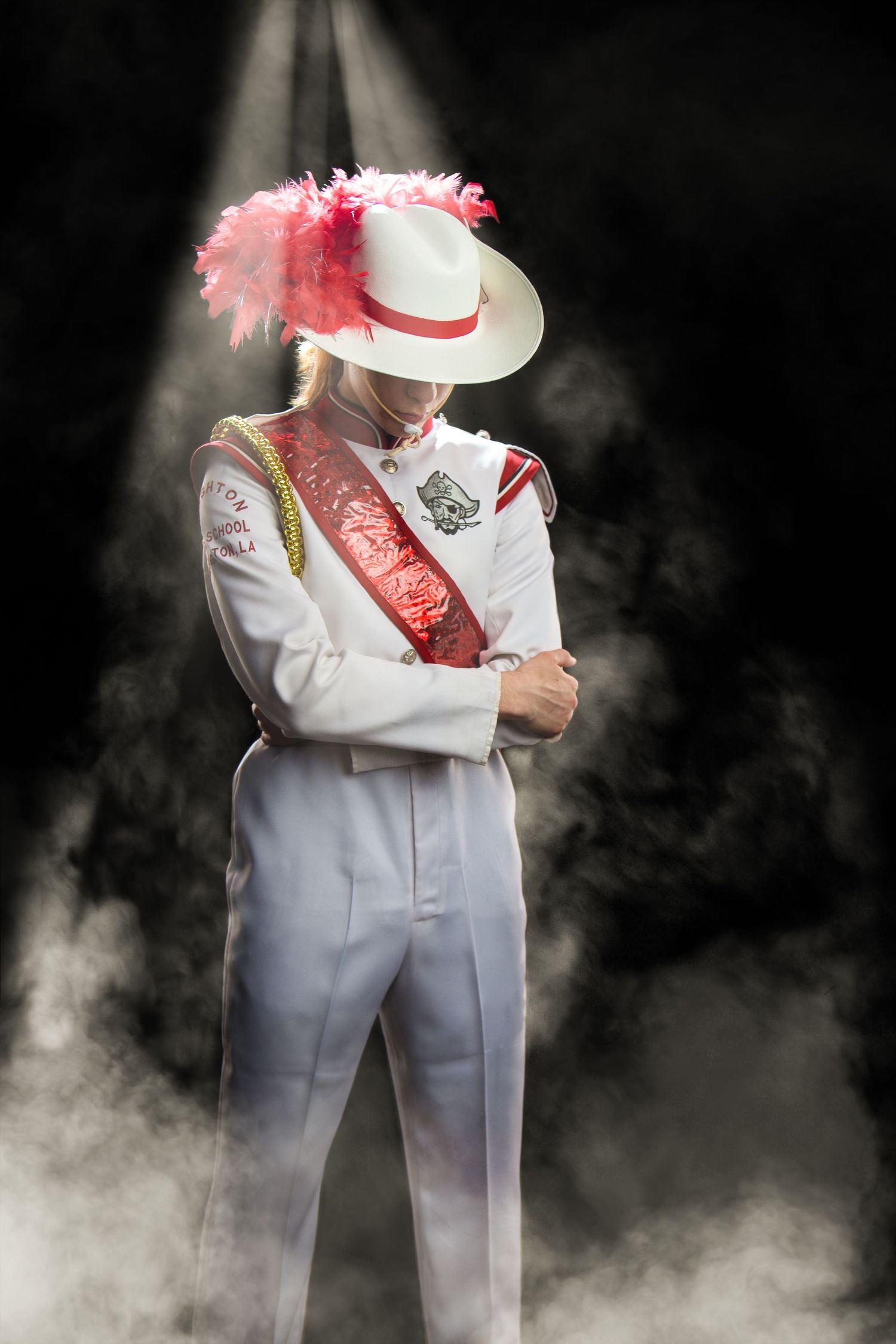 A figure in a white suit and pink feathered hat stands dramatically in fog against a dark background.