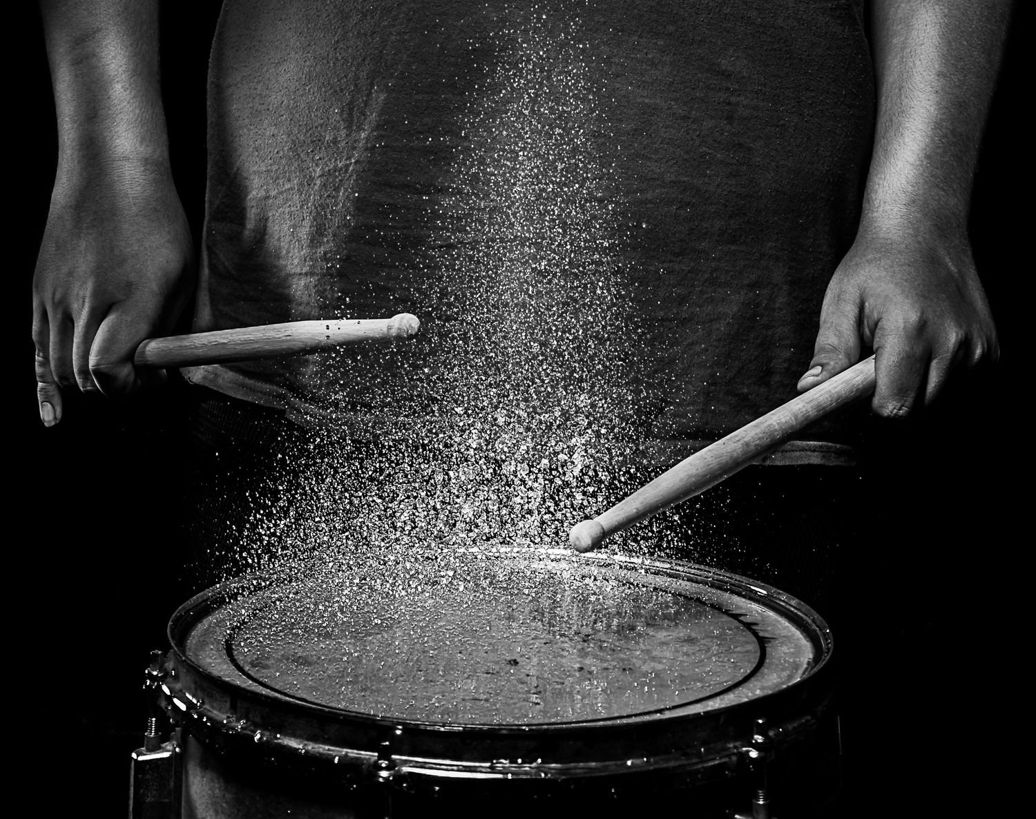 A drummer's hands are captured in black and white while playing a snare drum with powder or dust flying off the surface.