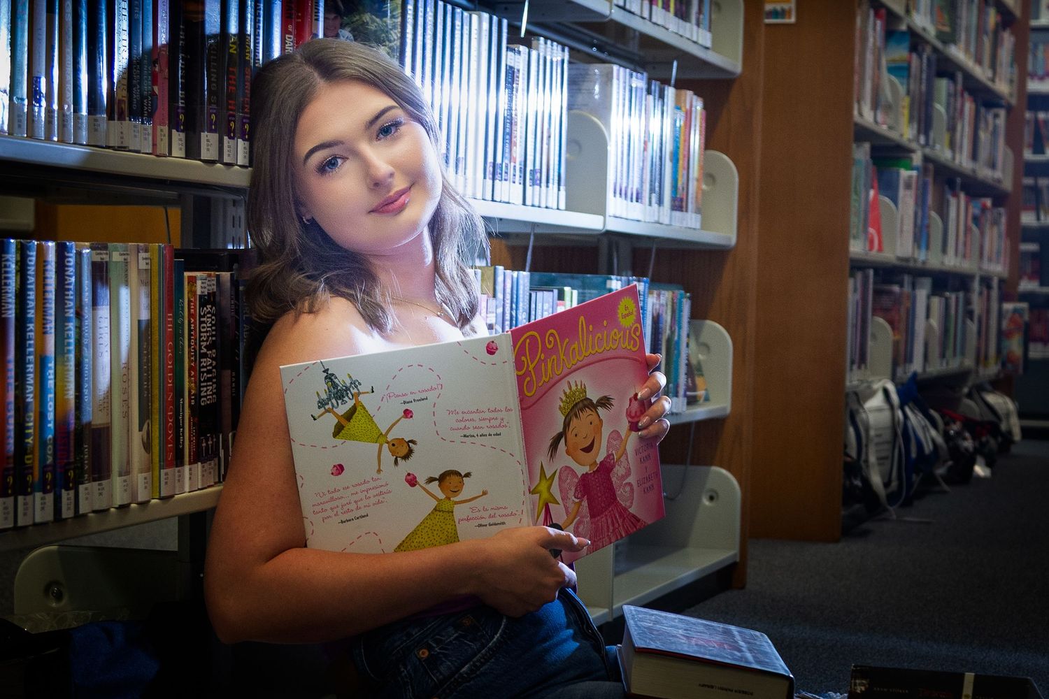 A person reads a children's book while sitting in a library with bookshelves visible in the background.