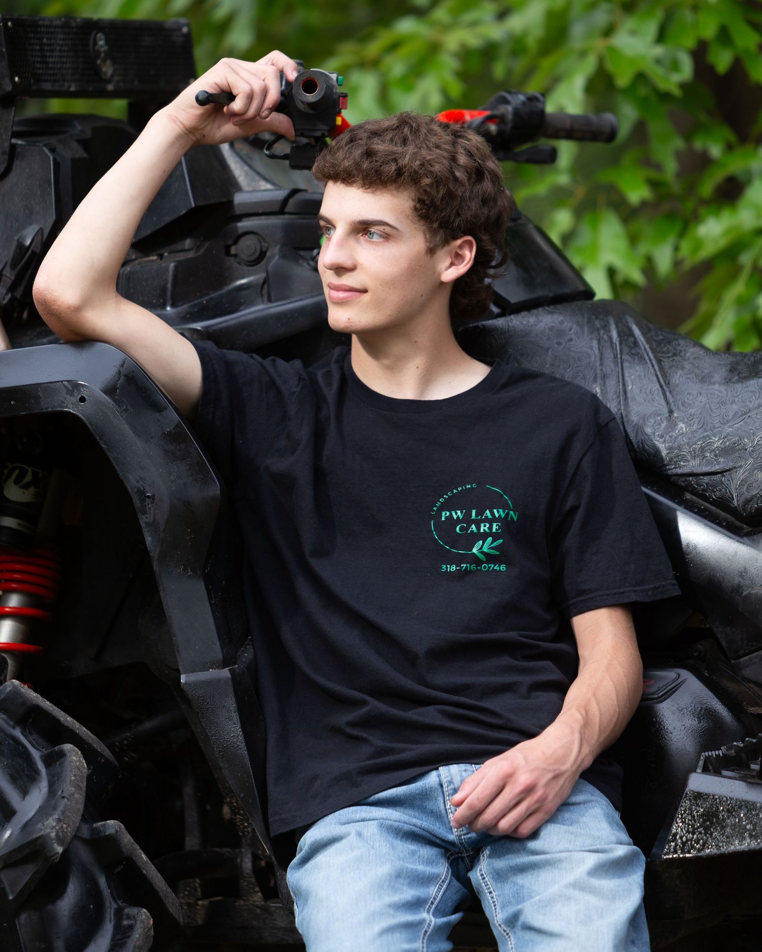 A person wearing a black t-shirt and jeans sits on a black bench against a leafy green background.