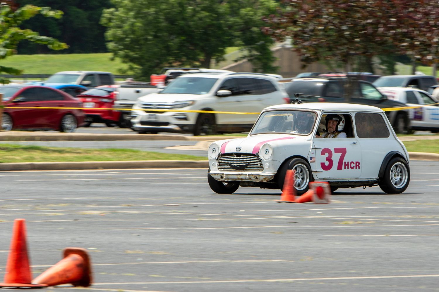 White Mini Cooper #37 competing in an autocross race, navigating orange cones in a parking lot during a sunny day.