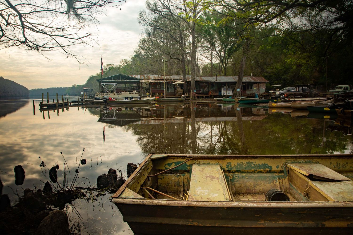An old weathered rowboat sits on a calm reflective lake near a rustic dock and trees at dusk.
