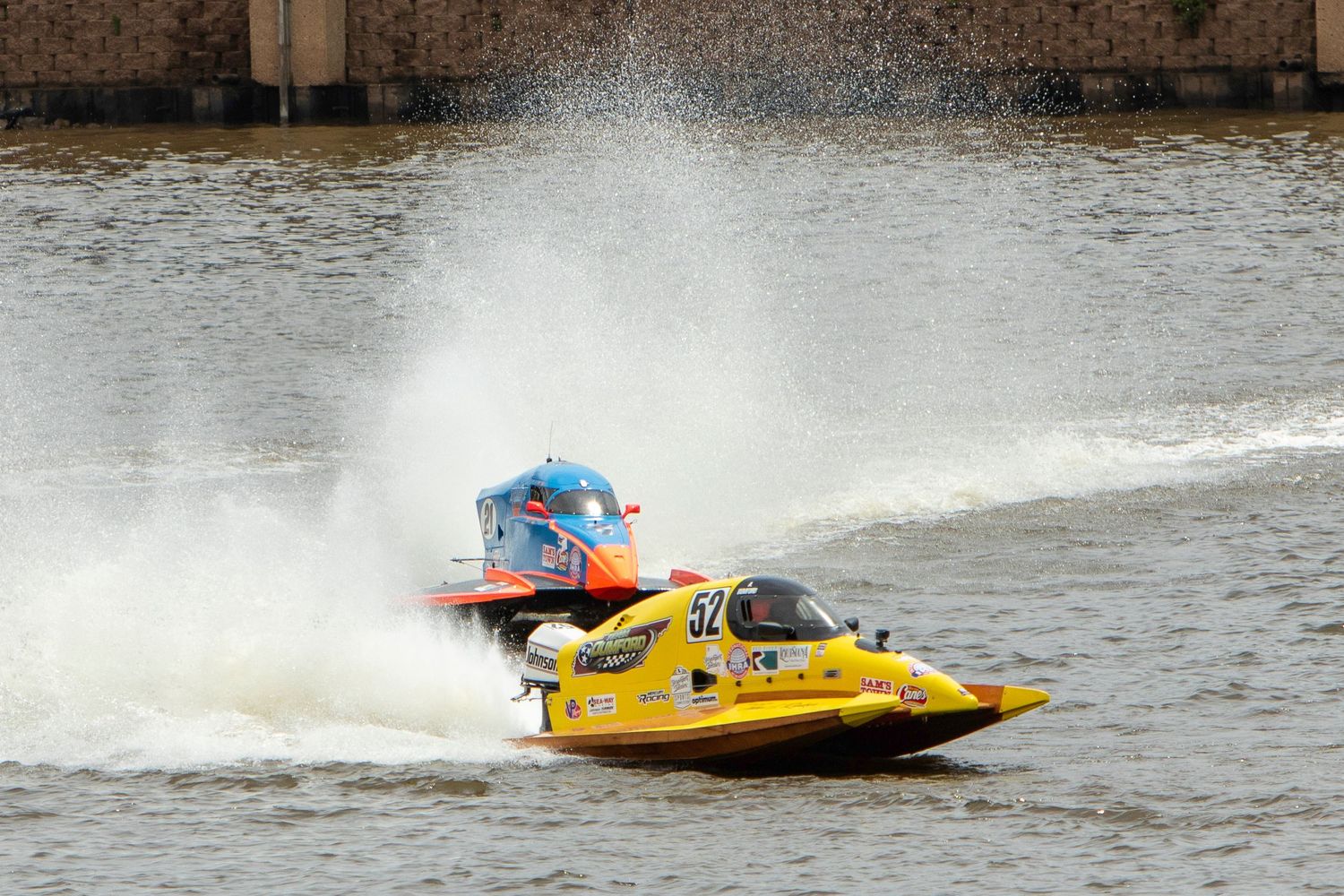 Two colorful speedboats racing on a river, with a yellow boat in the foreground and a blue boat behind, creating large water sprays.