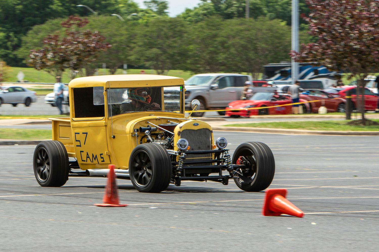 Yellow vintage hot rod car number 57 navigating orange cones at an autocross event in a parking lot.