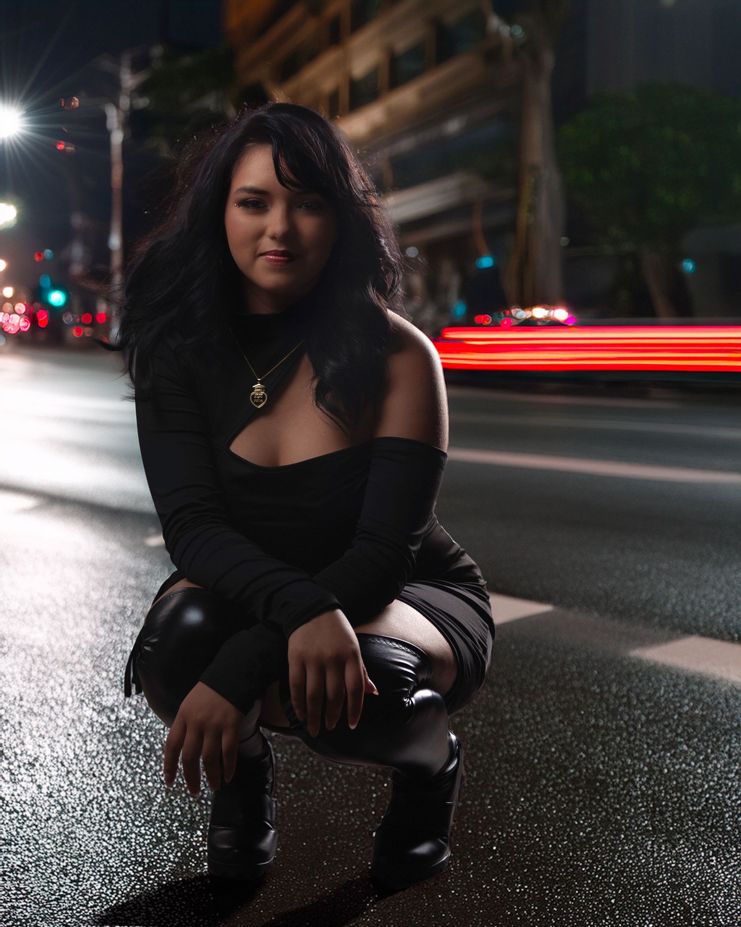 A person in black clothing crouches on a wet asphalt road at night with blurred lights in the background.