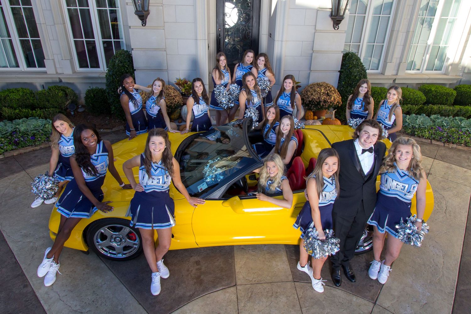 Group of cheerleaders in blue uniforms posing around a yellow sports car in front of an elegant mansion.