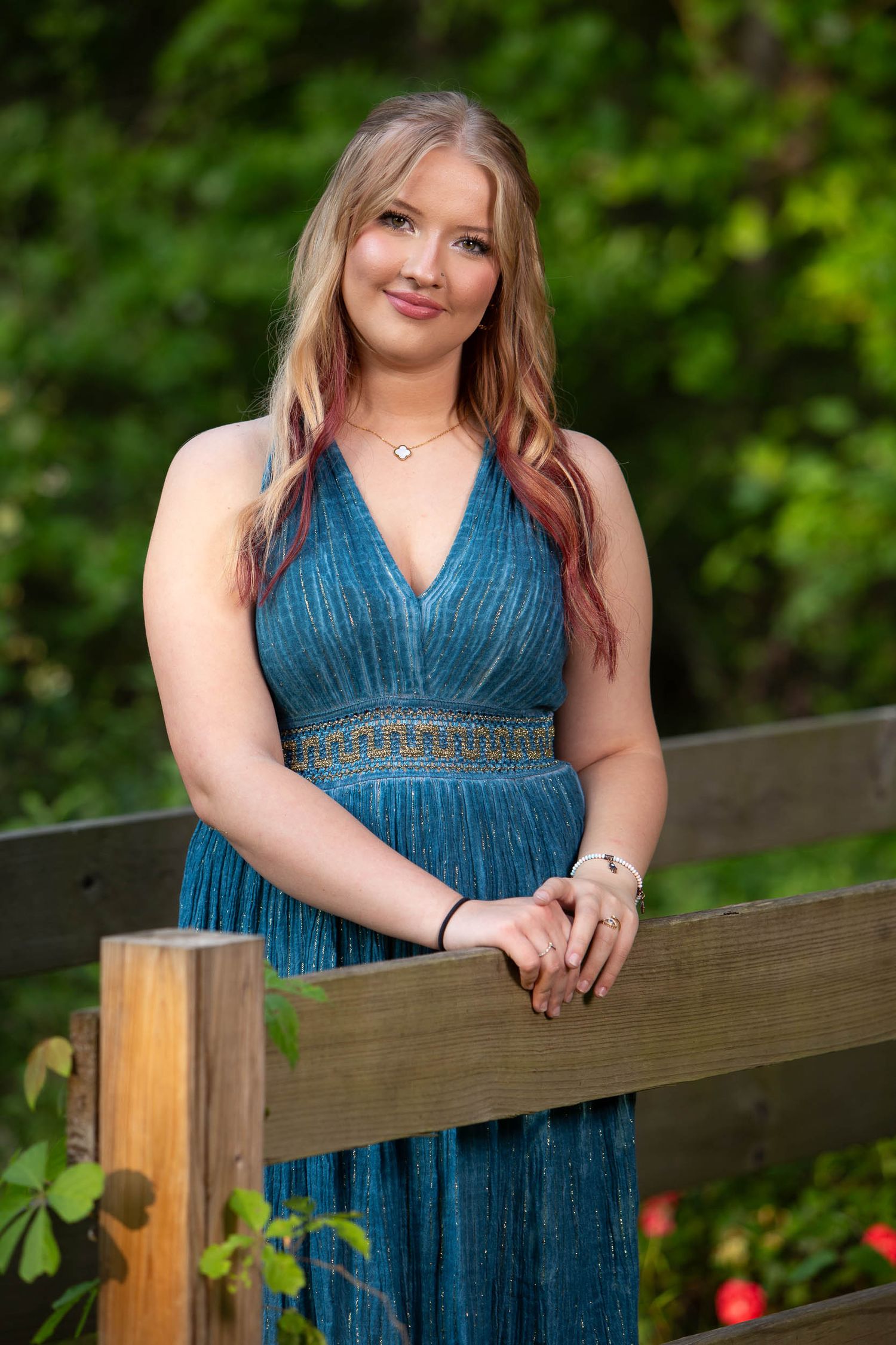 Someone wearing a blue evening gown leans against a wooden railing with greenery in the background.