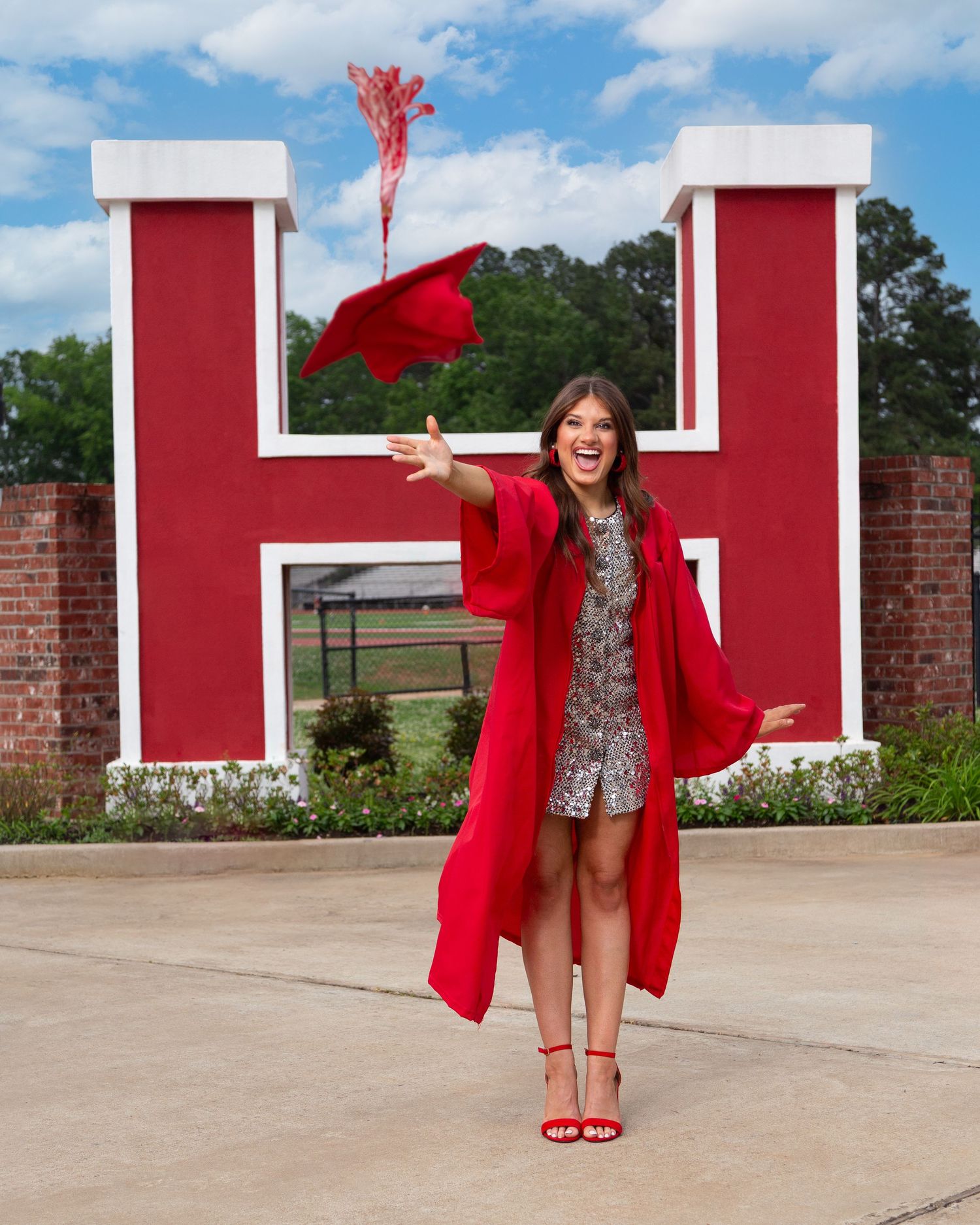 A person in red heels and coat poses in front of large red letters against a brick wall on a sunny day.