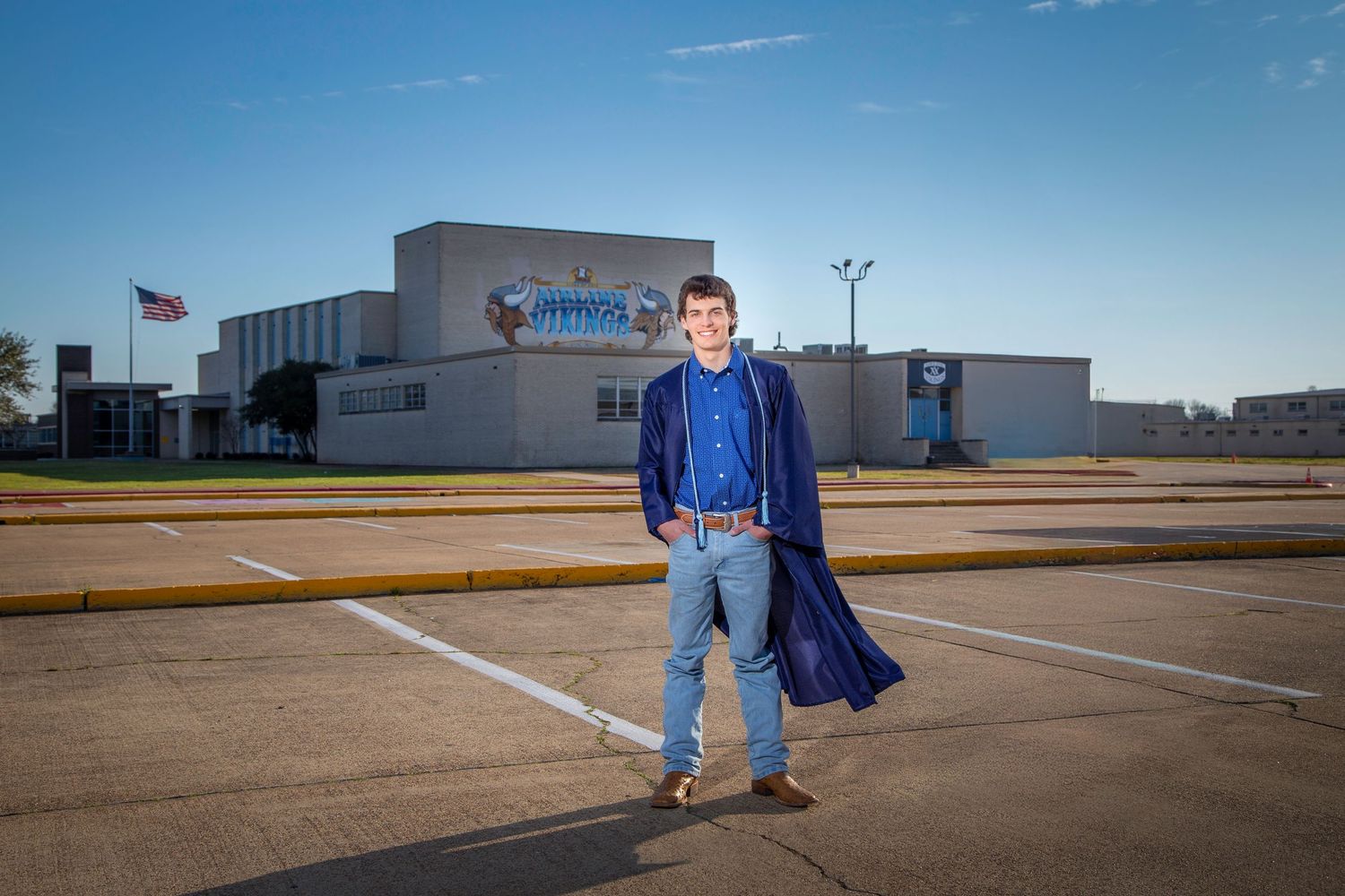A male high school senior in a blue cape stands in an empty baseball field at Airline high school on a sunny day with clear blue skies.