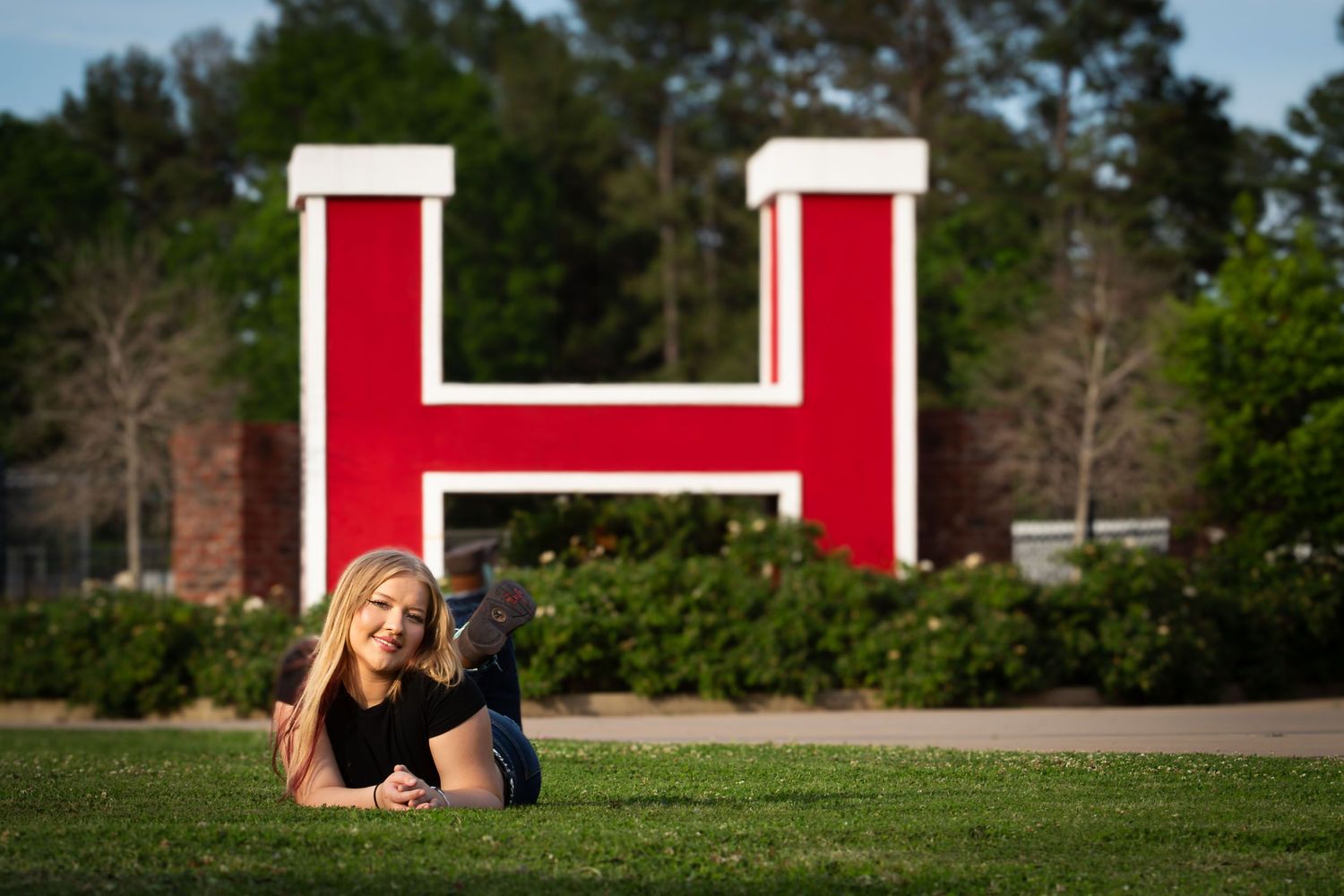 Student reading while lying on grass near red and white campus buildings on a sunny day.