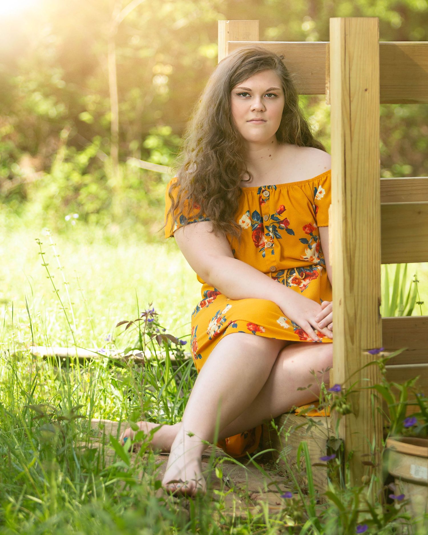 Young woman in orange floral off-shoulder dress sitting outdoors on wooden chair surrounded by lush green grass and wildflowers.
