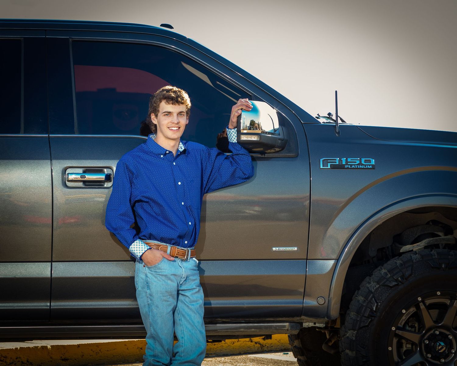 A person in a blue shirt and jeans stands next to a dark gray pickup truck in a parking lot.