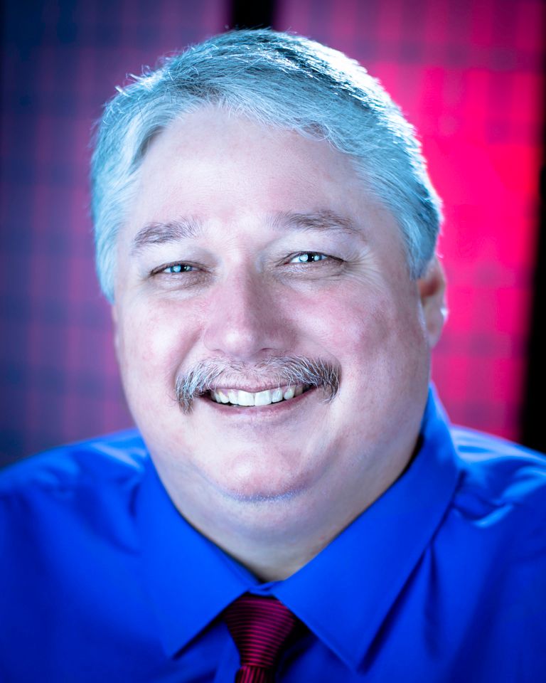 A  headshot showing Shreveport photographer in a blue shirt and red tie with smiling expression against a red background.
