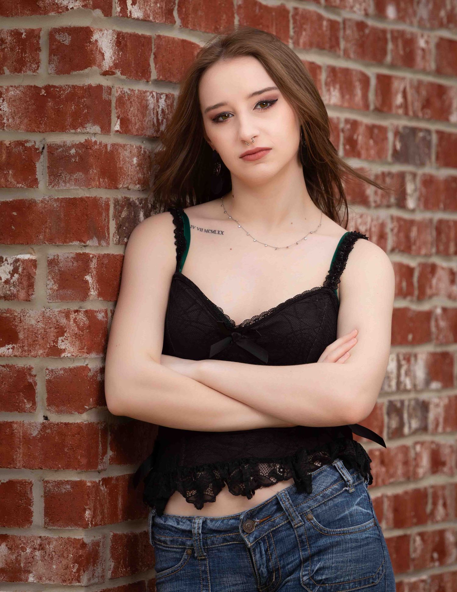 Young woman in black lace top and jeans poses against rustic brick wall in artistic portrait series.