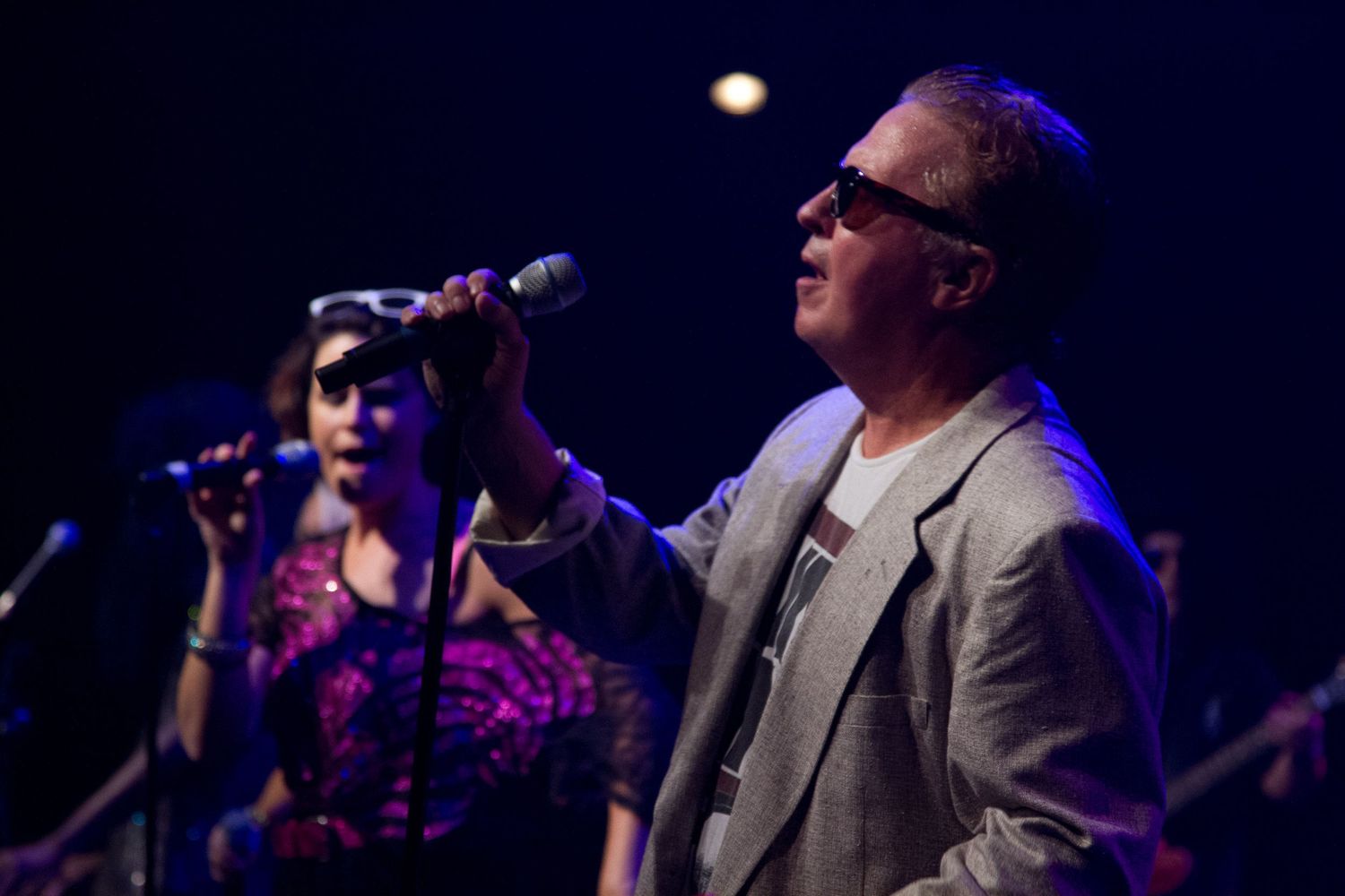 Two singers performing on stage, man in gray blazer and sunglasses holding microphone with woman vocalist behind him.