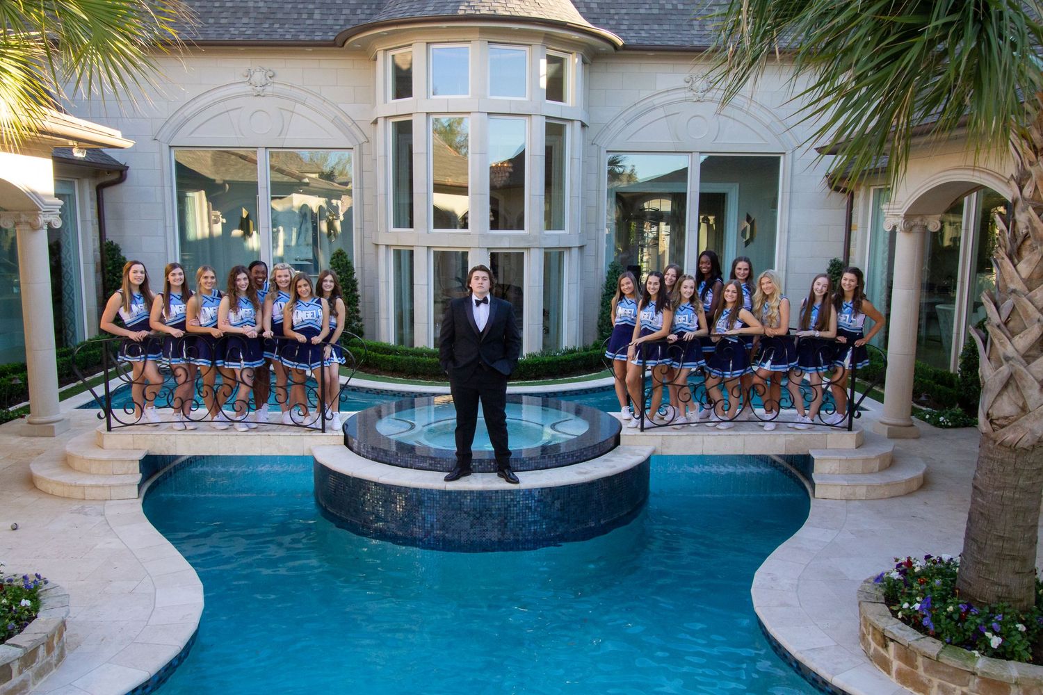 A groom in a tuxedo stands by a luxury pool surrounded by bridesmaids in blue dresses at an elegant mansion.