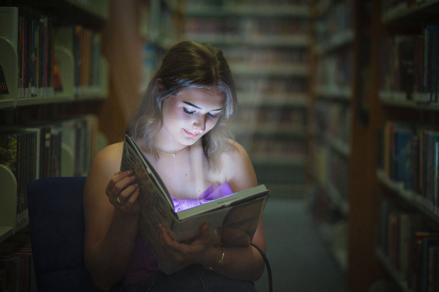 A student reads illuminated by digital tablet light while studying late at night in a library between bookshelves.