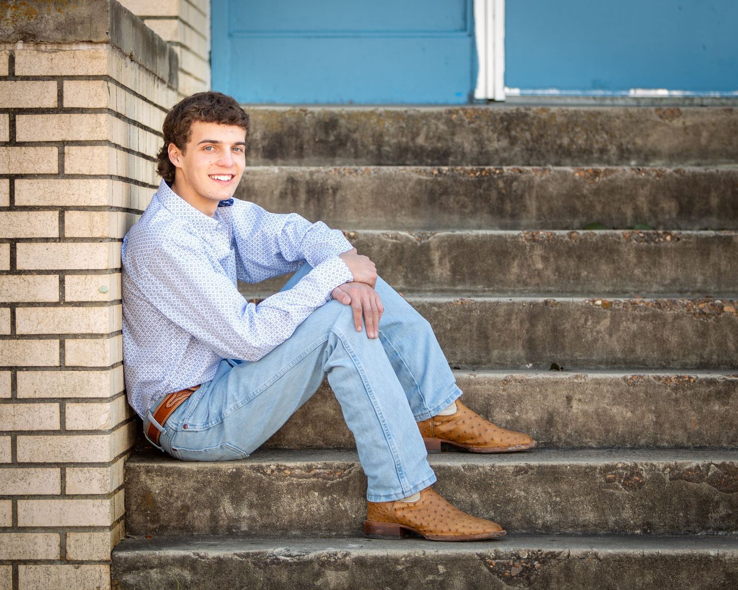 A person in light blue jeans and brown boots reclines casually on concrete steps next to a tan brick wall.