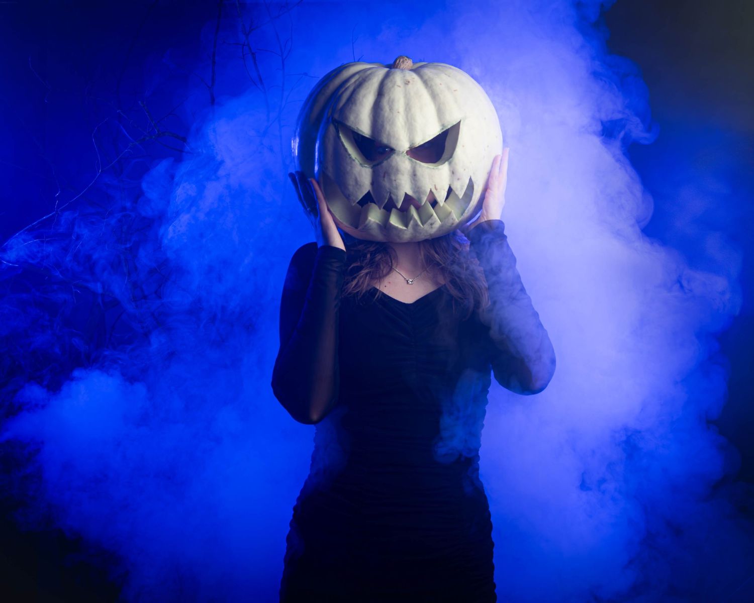 A figure in a pumpkin head mask stands mysteriously in blue fog creating an eerie Halloween atmosphere.