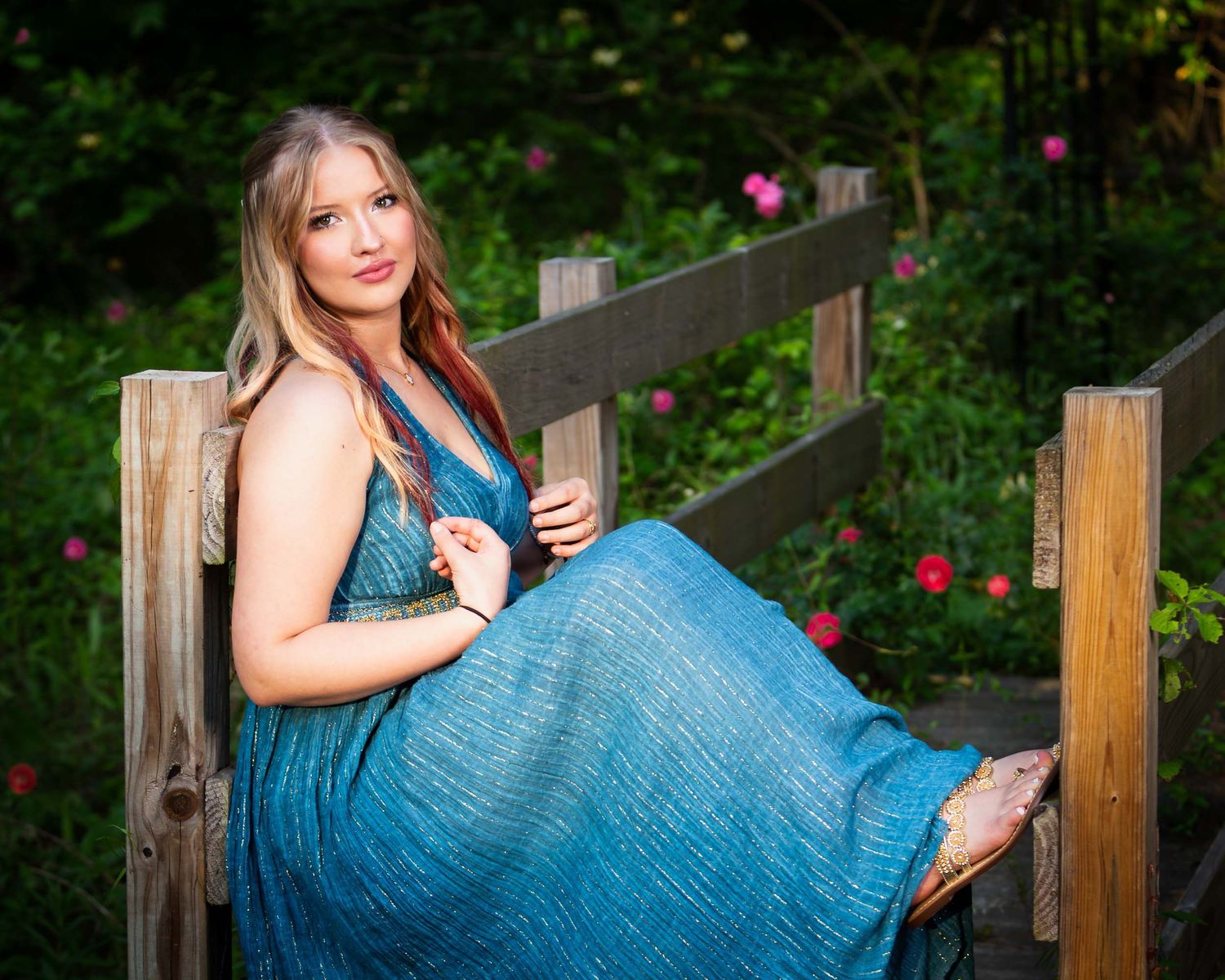 A person in a flowing blue sequined dress poses outdoors against a wooden fence in a garden setting.