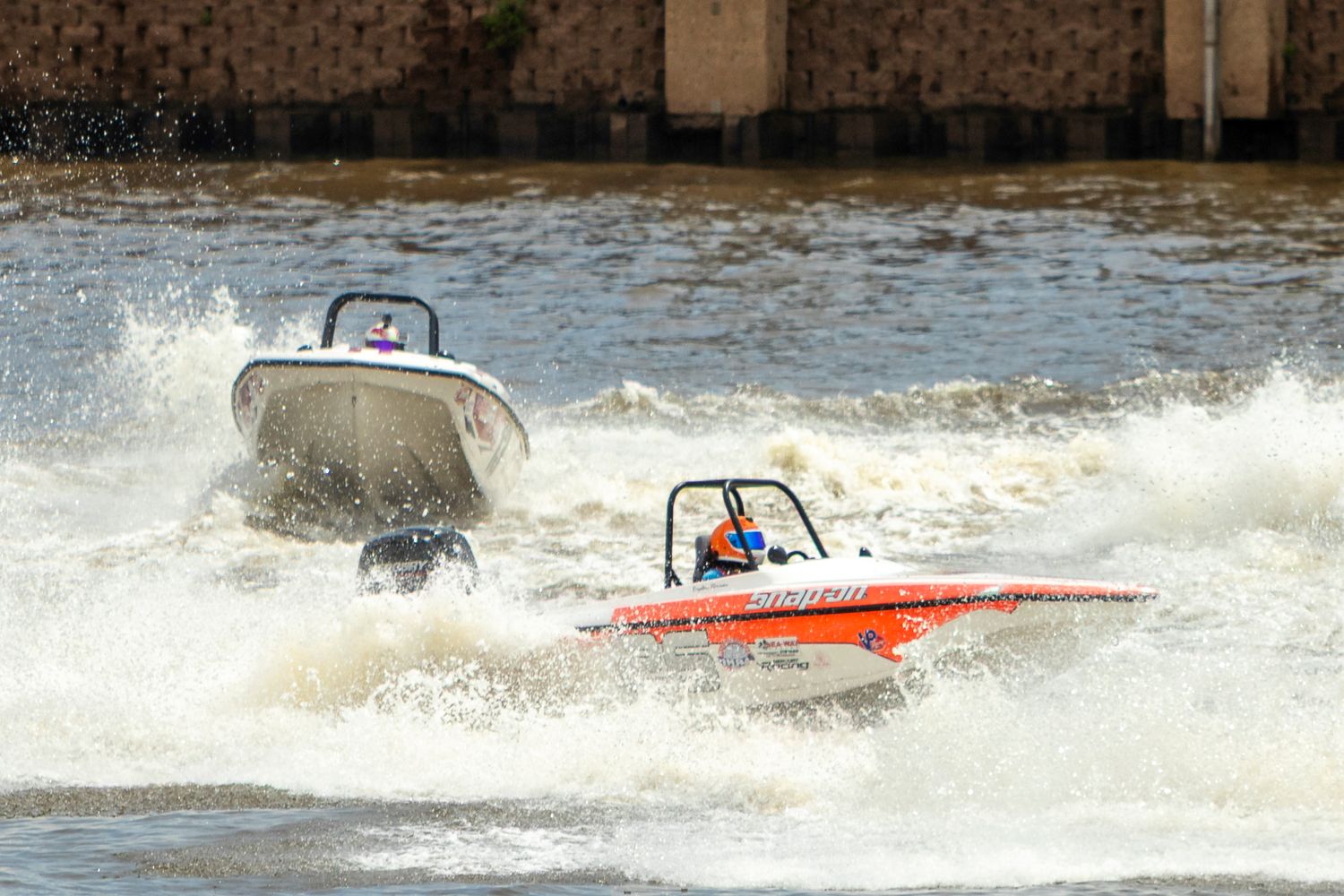 Two speedboats racing on choppy water, an orange boat in front and a white boat close behind, creating large splashing waves.