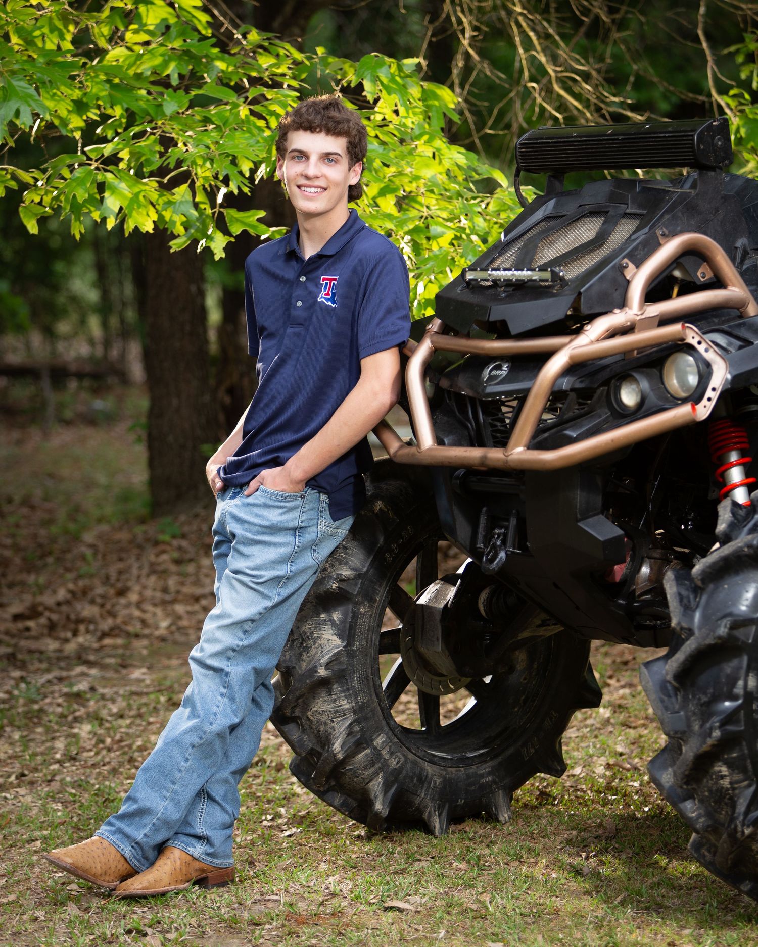 A person in jeans and a navy shirt poses casually next to a large tractor tire in an outdoor setting.