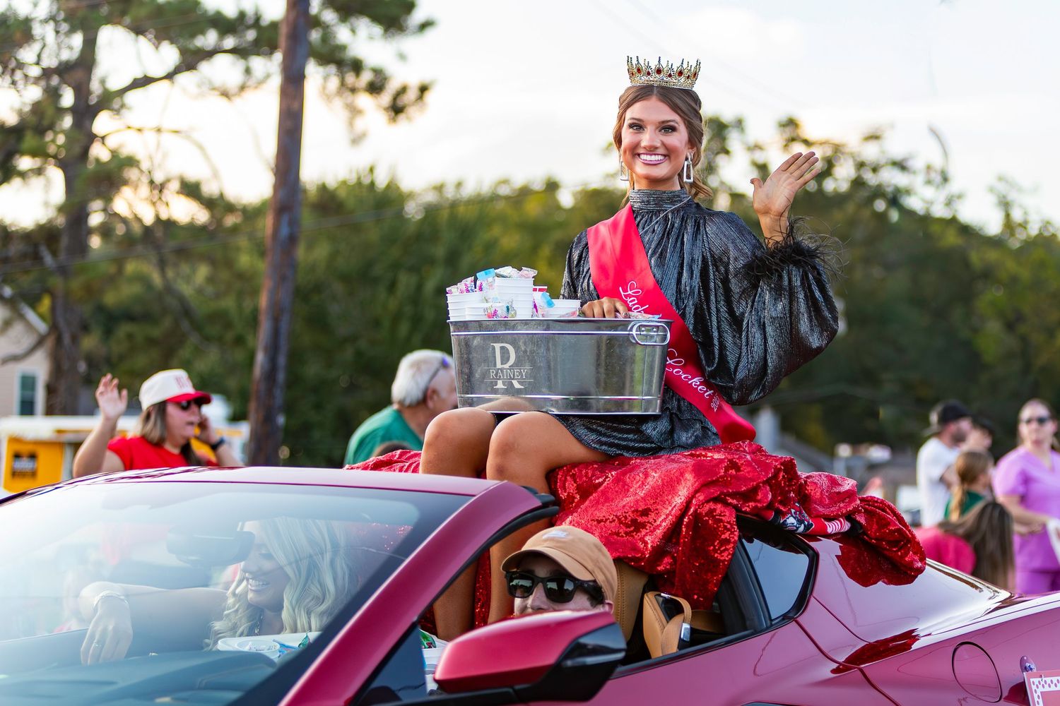 Young woman in graduation gown and crown waves from pink convertible during homecoming parade.