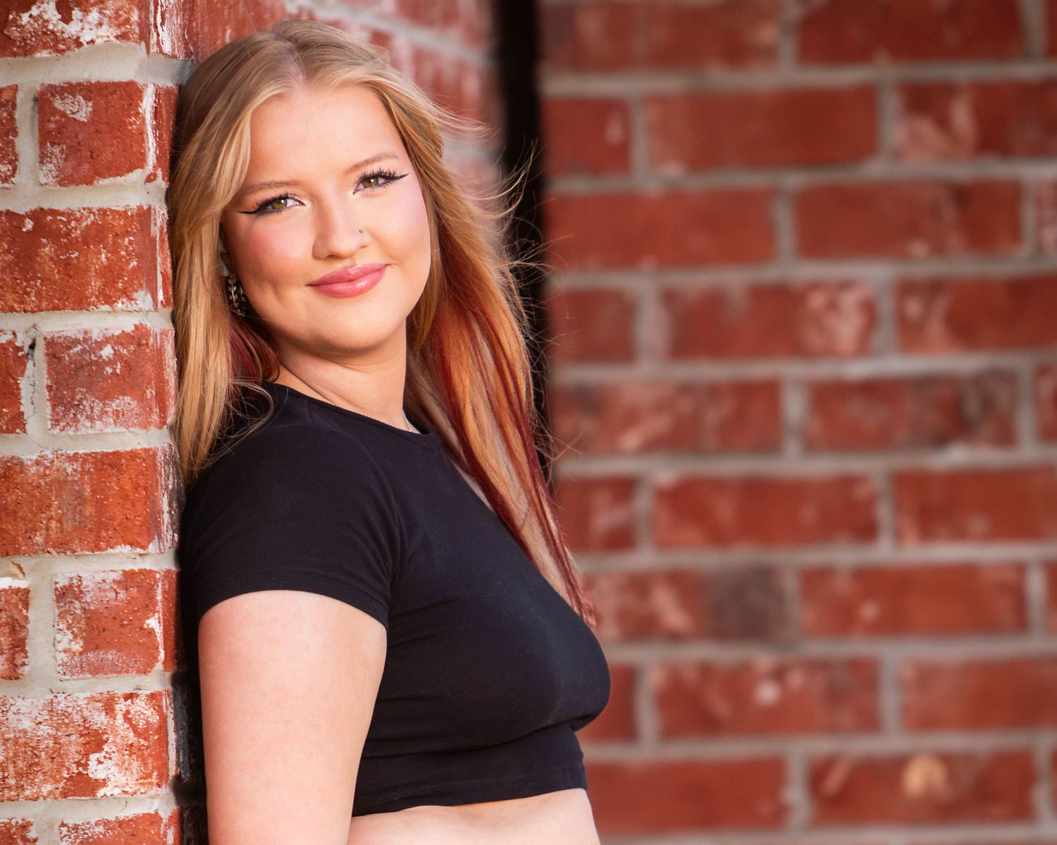 high school senior in black top poses against rustic red brick wall background in professional portrait setting.