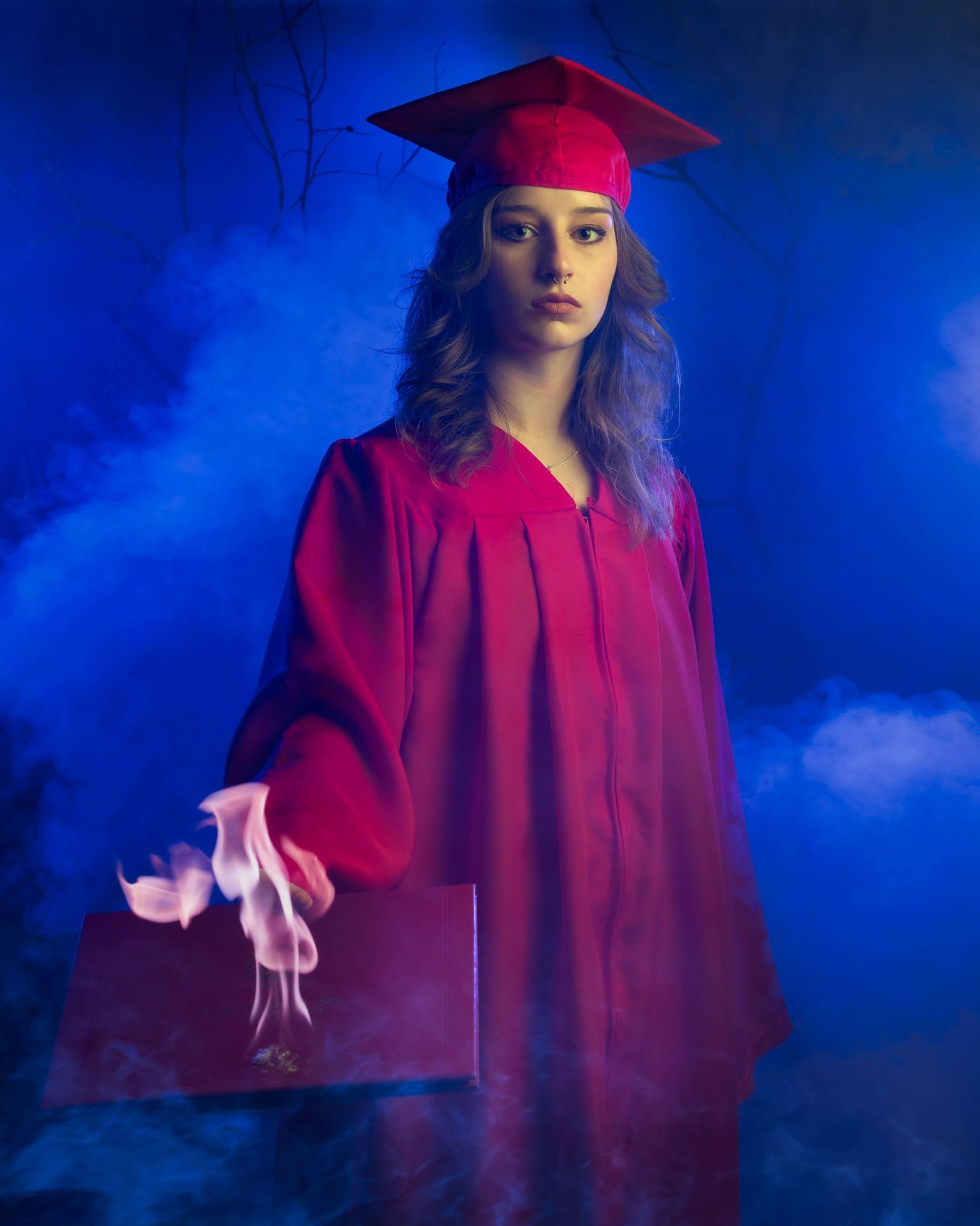 Graduate in red cap and gown poses against dramatic blue backdrop with artistic lighting effects.