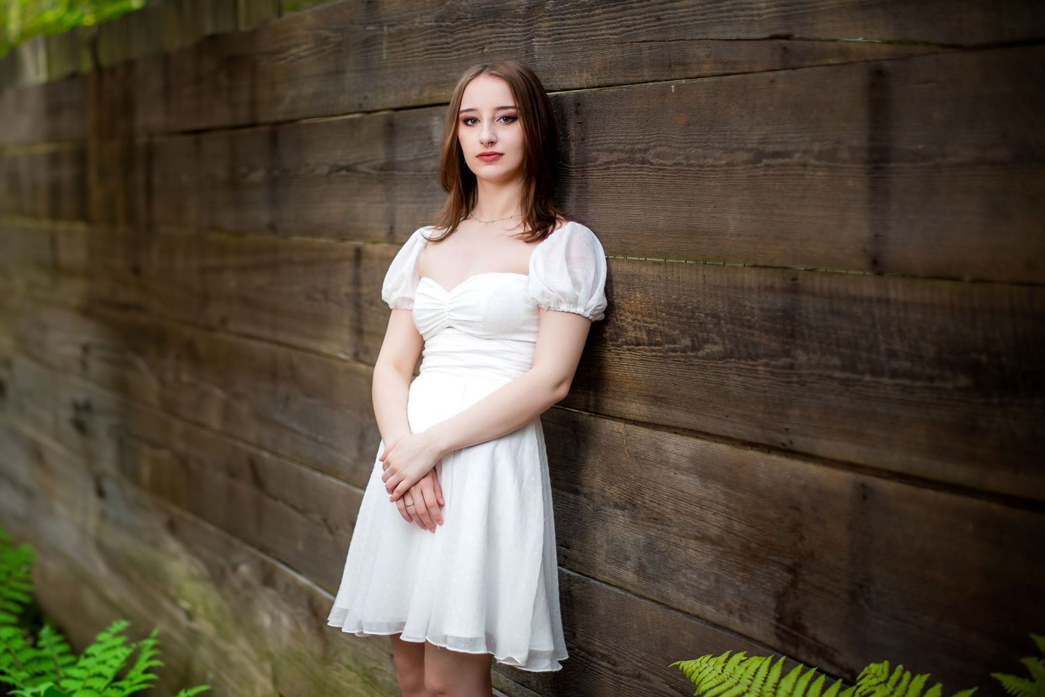 A high school senior in a white dress poses against a rustic wooden wall with green ferns in the foreground.