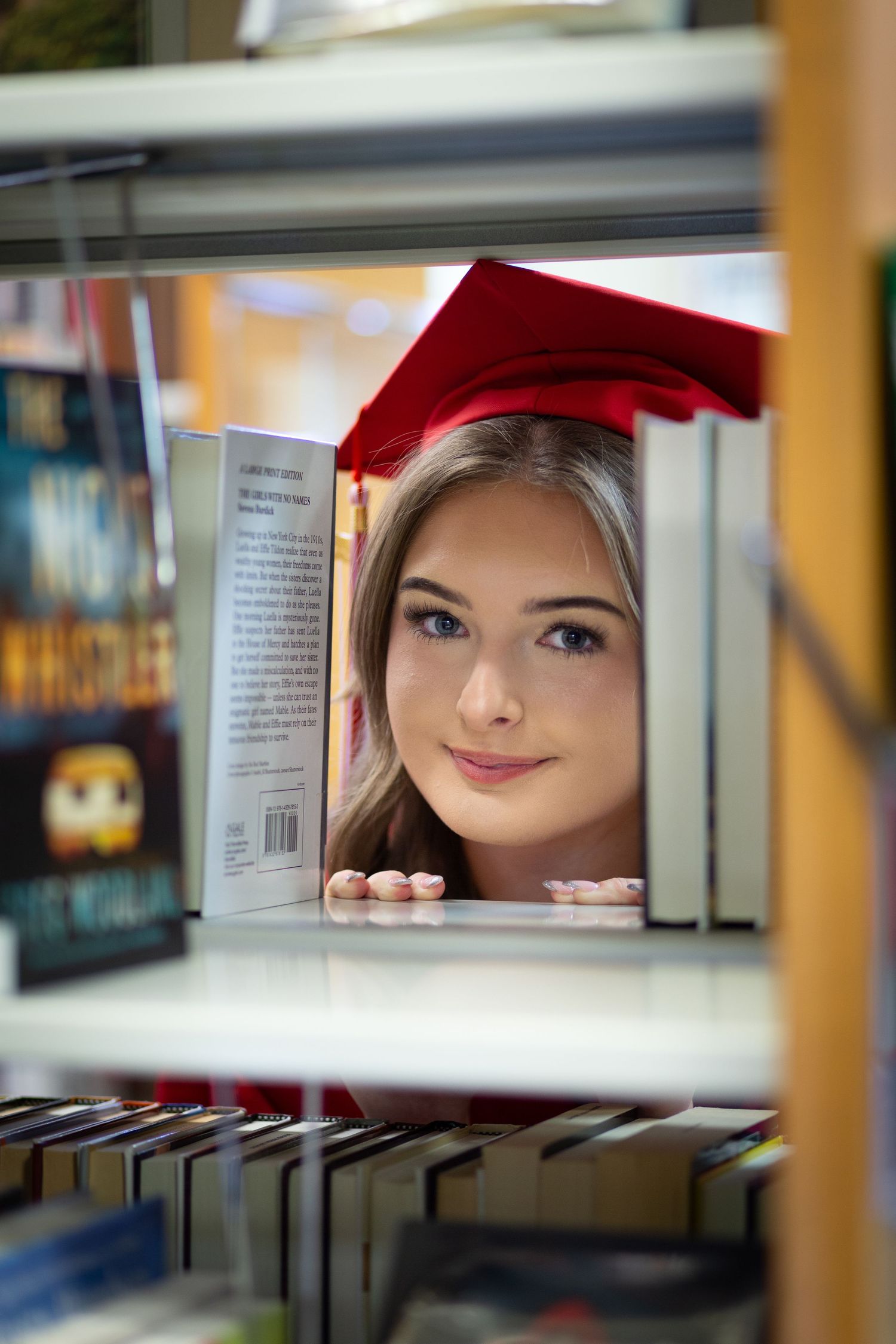 A creative perspective shot through library shelves showing glimpses of a person in a red graduation cap.