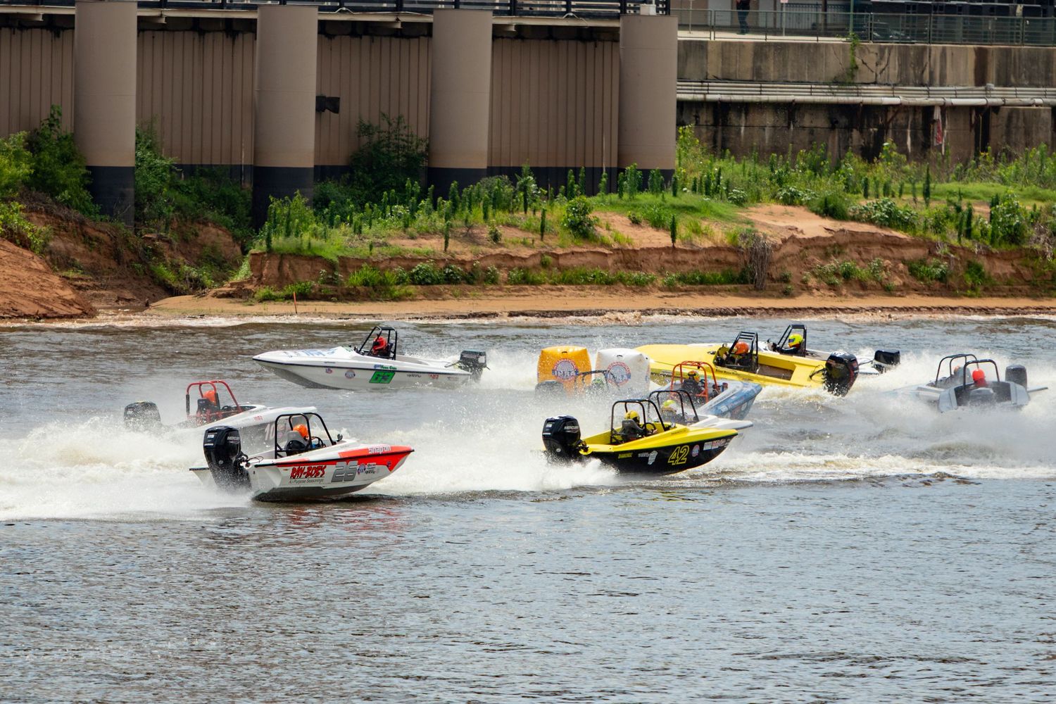 Multiple colorful speedboats racing across a river near a concrete bridge during a competitive powerboat racing event.