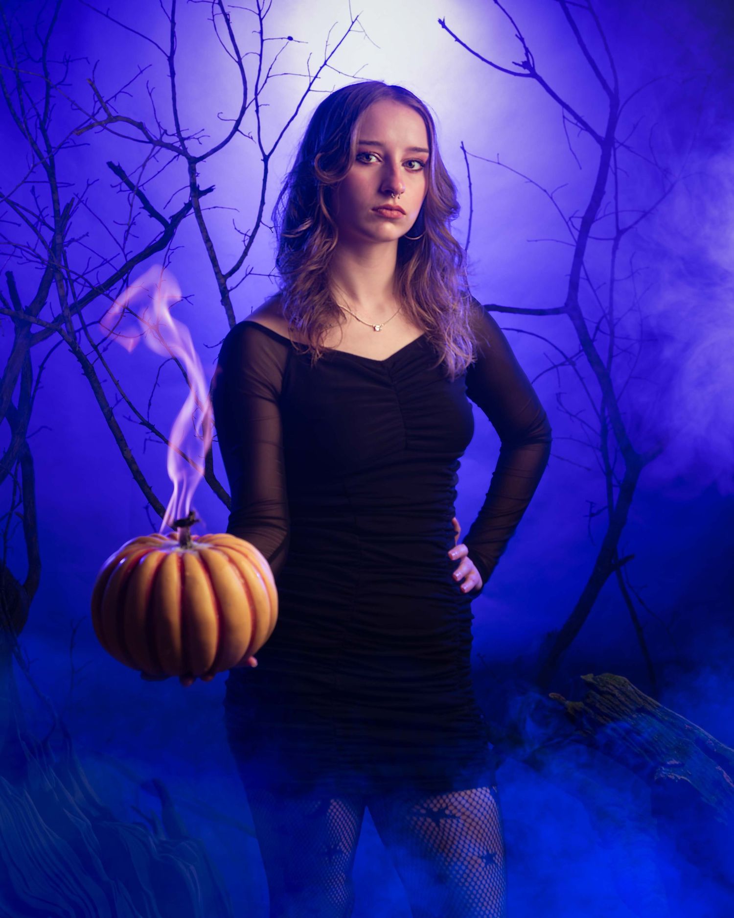 Atmospheric Halloween portrait with black dress against purple fog and spooky trees, featuring glowing pumpkin prop.